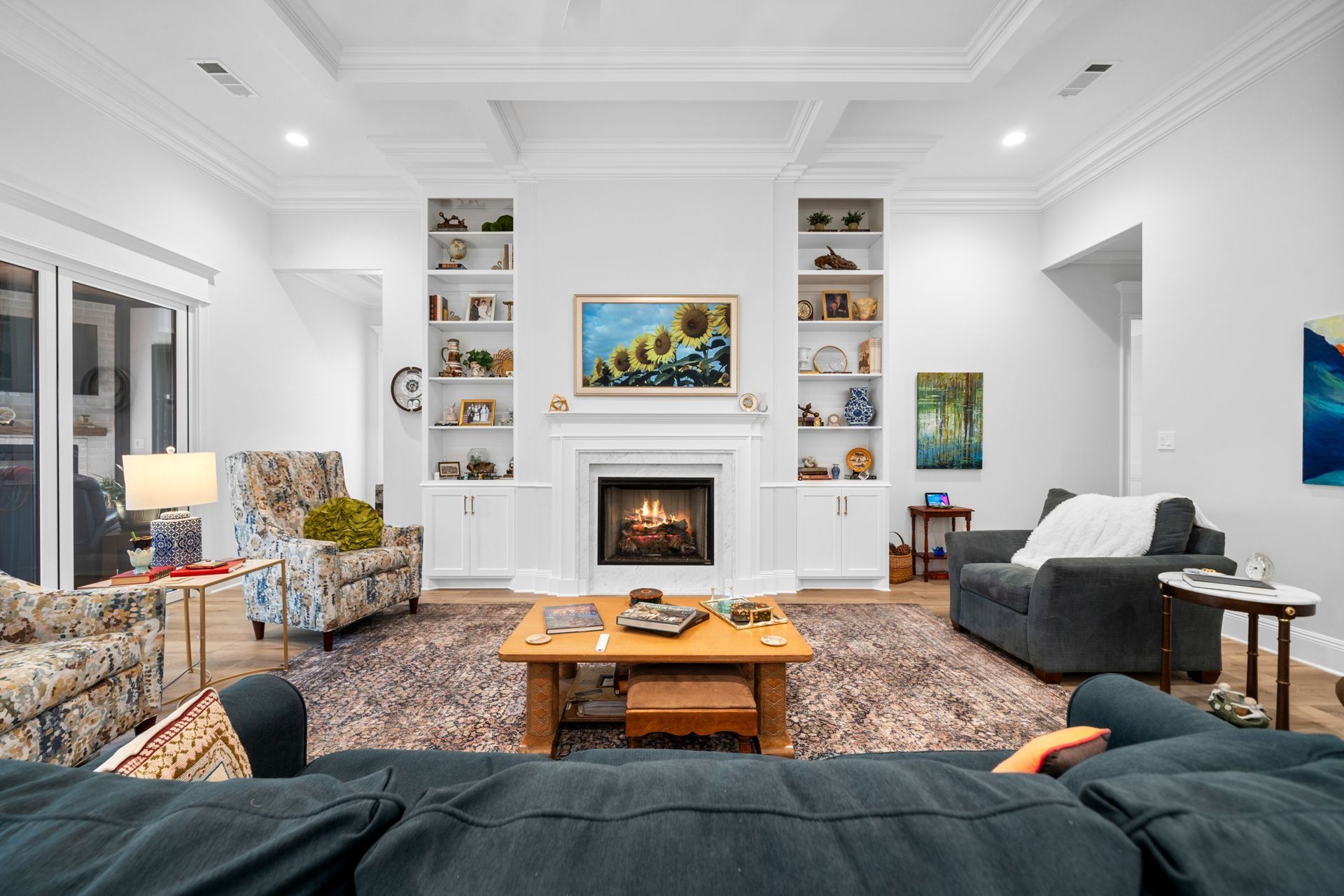 A living room with a fireplace, built-in shelves, patterned armchairs, a gray sofa, and a wooden coffee table on a rug.