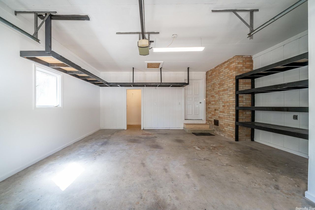 An empty garage with a concrete floor, overhead metal storage racks, white walls, and a stone accent wall with shelving.