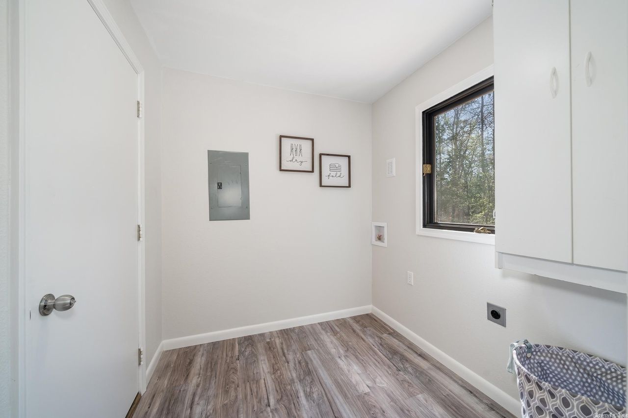 A laundry room with light gray walls, wood-look flooring, a window, wall-mounted cabinets, and a electrical panel.