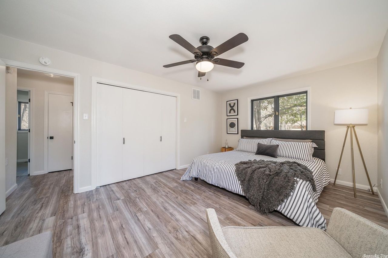 A bright bedroom with wood-look flooring, a queen bed with striped bedding, white sliding closets, and a tripod floor lamp.