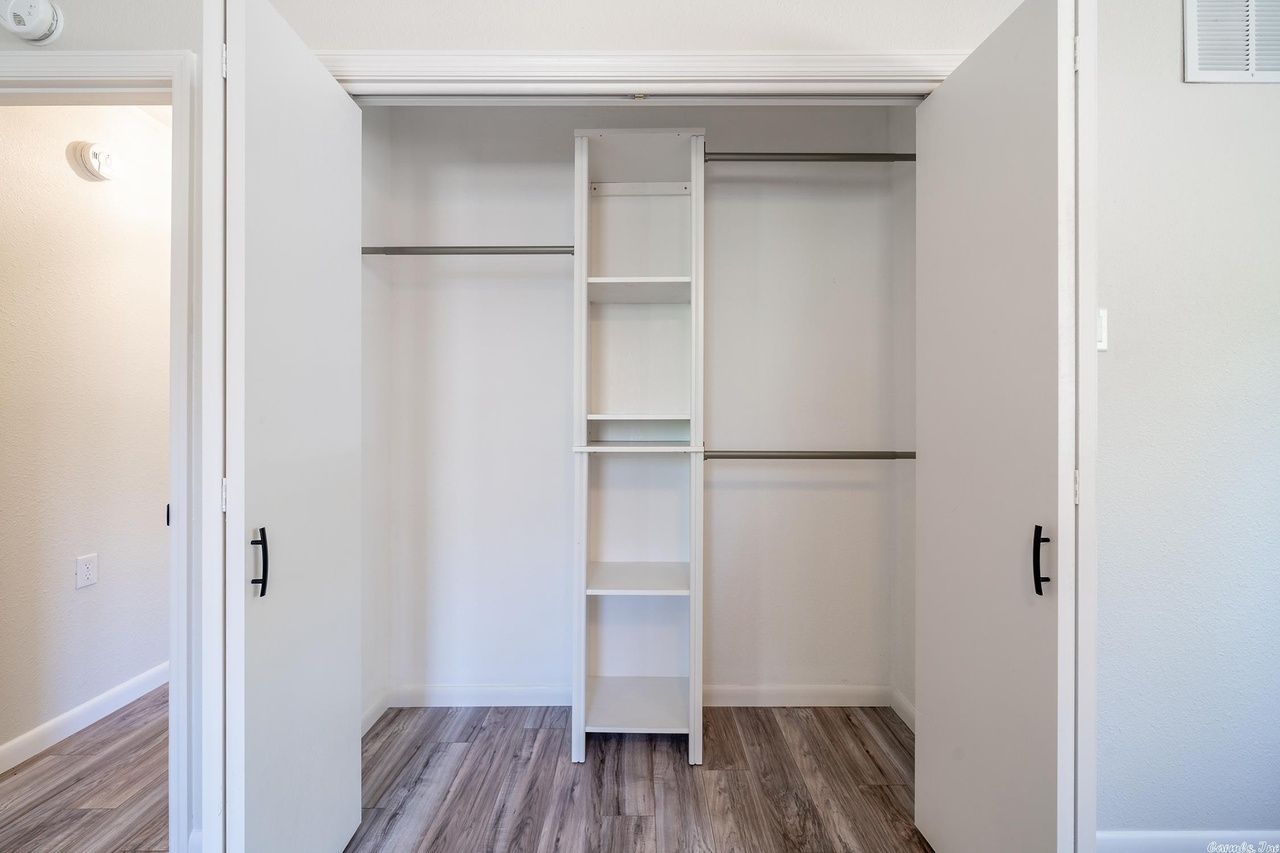 A white reach-in closet with central shelving and two hanging rods, set against light gray walls and wood-pattern flooring.