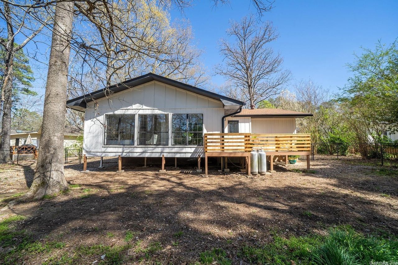 A white, single-story house with a wooden deck stands among trees on a sunny day.