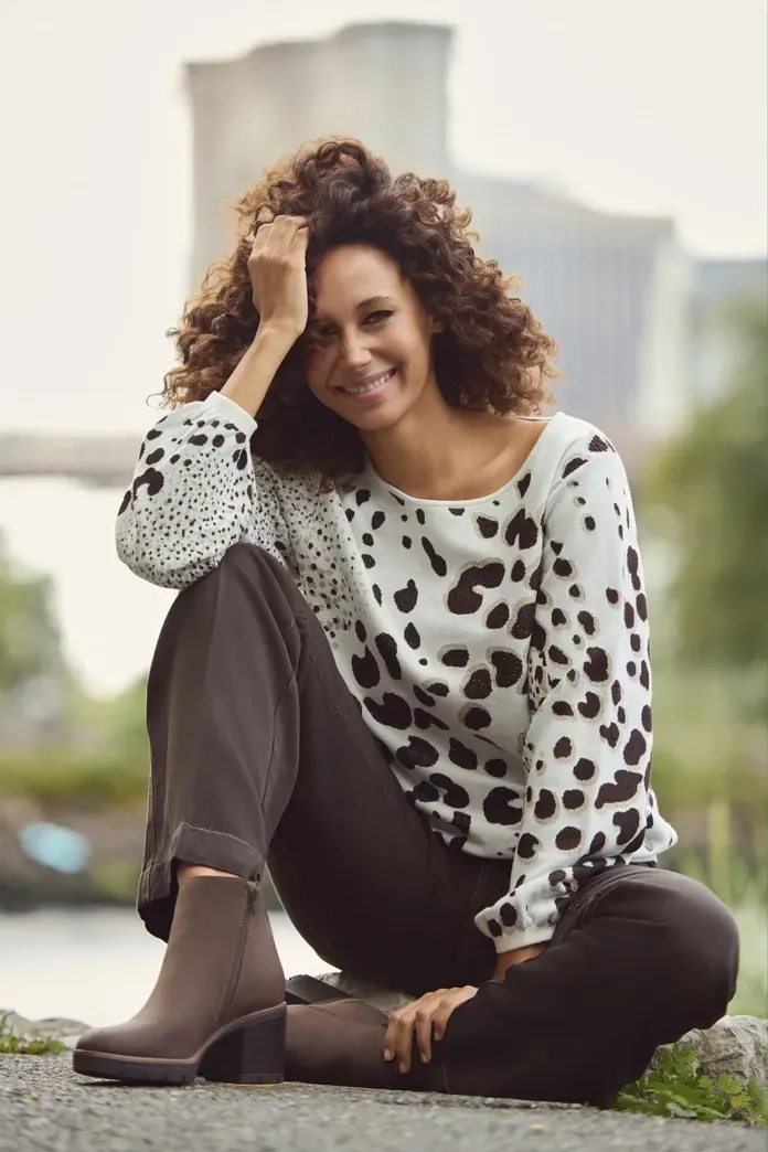 Frau in gepunktetem Pullover und brauner Hose, lächelnd, sitzt im Freien.