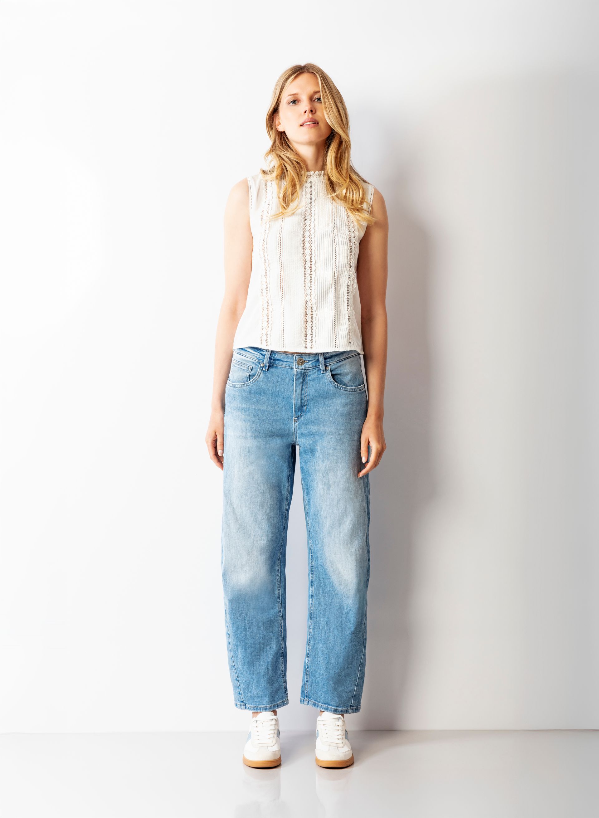 Frau in einem ärmellosen weißen Top, hellblauen Jeans und weißen Turnschuhen vor einer weißen Wand.