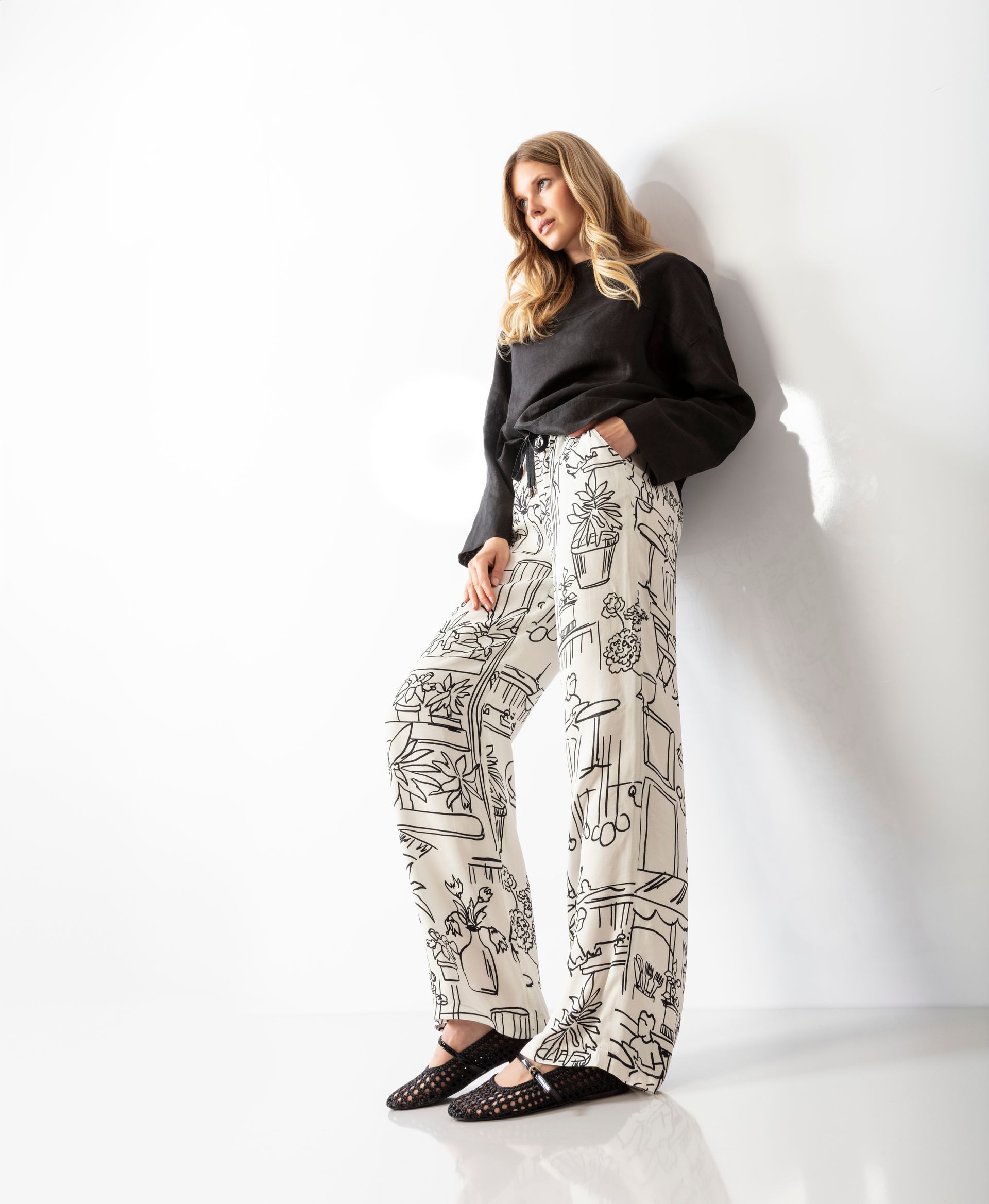 Eine Frau in schwarzem Oberteil und gemusterter Hose lehnt an einer weißen Wand.