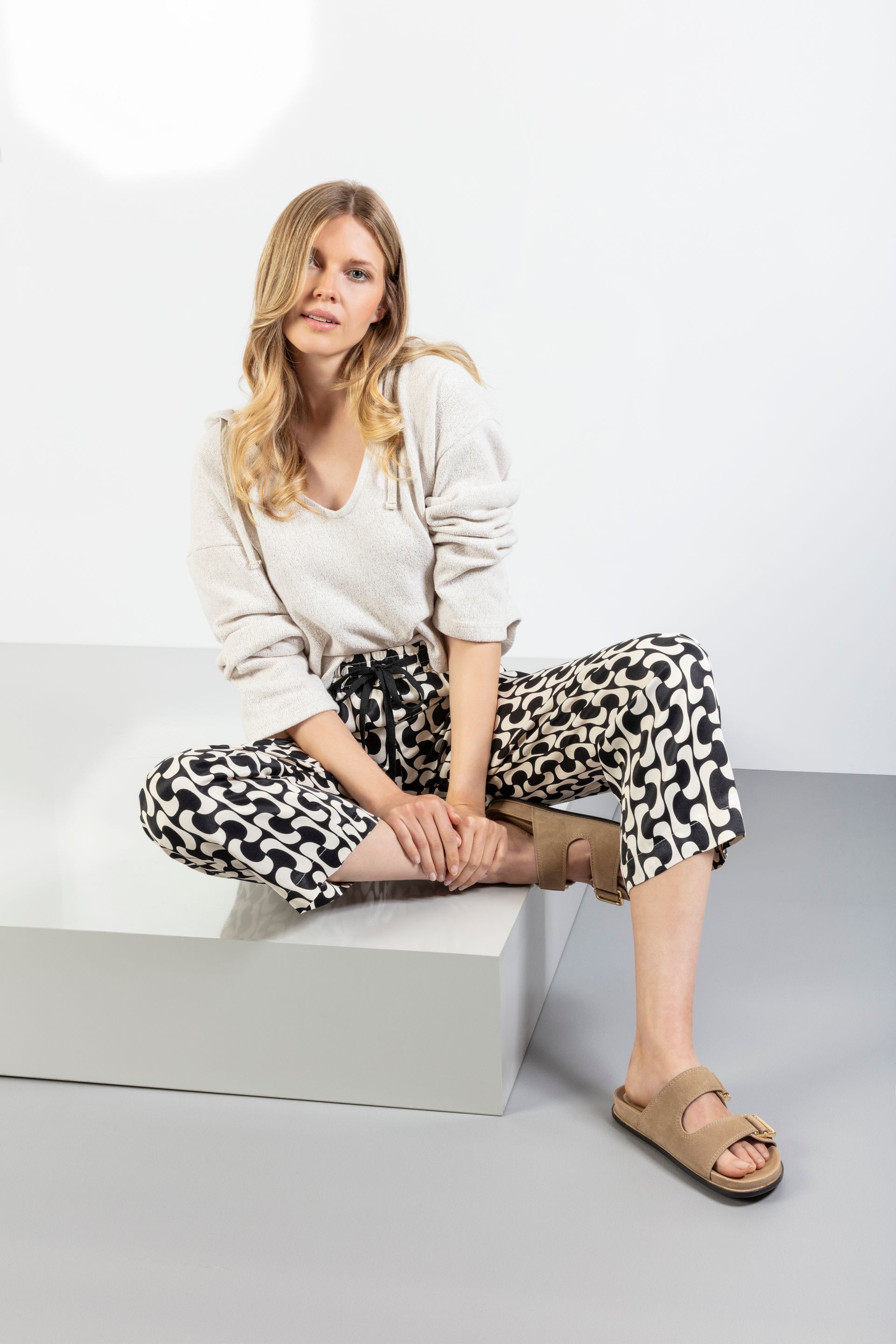 Eine Frau in Pullover und gemusterter Hose sitzt mit Sandalen auf einem grauen Block.