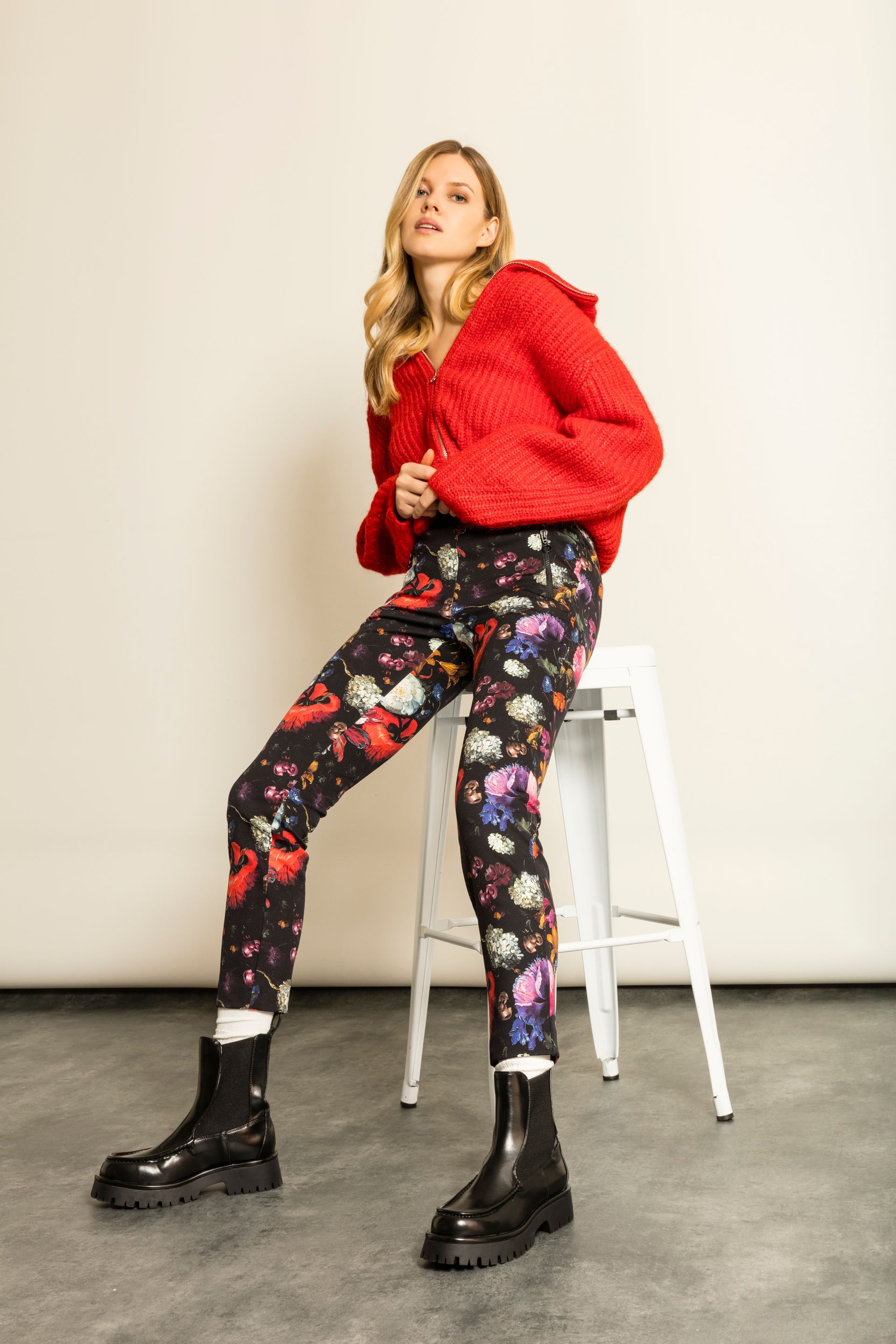 Eine Frau in roter Jacke und geblümter Hose sitzt auf einem weißen Hocker und trägt schwarze Stiefel.