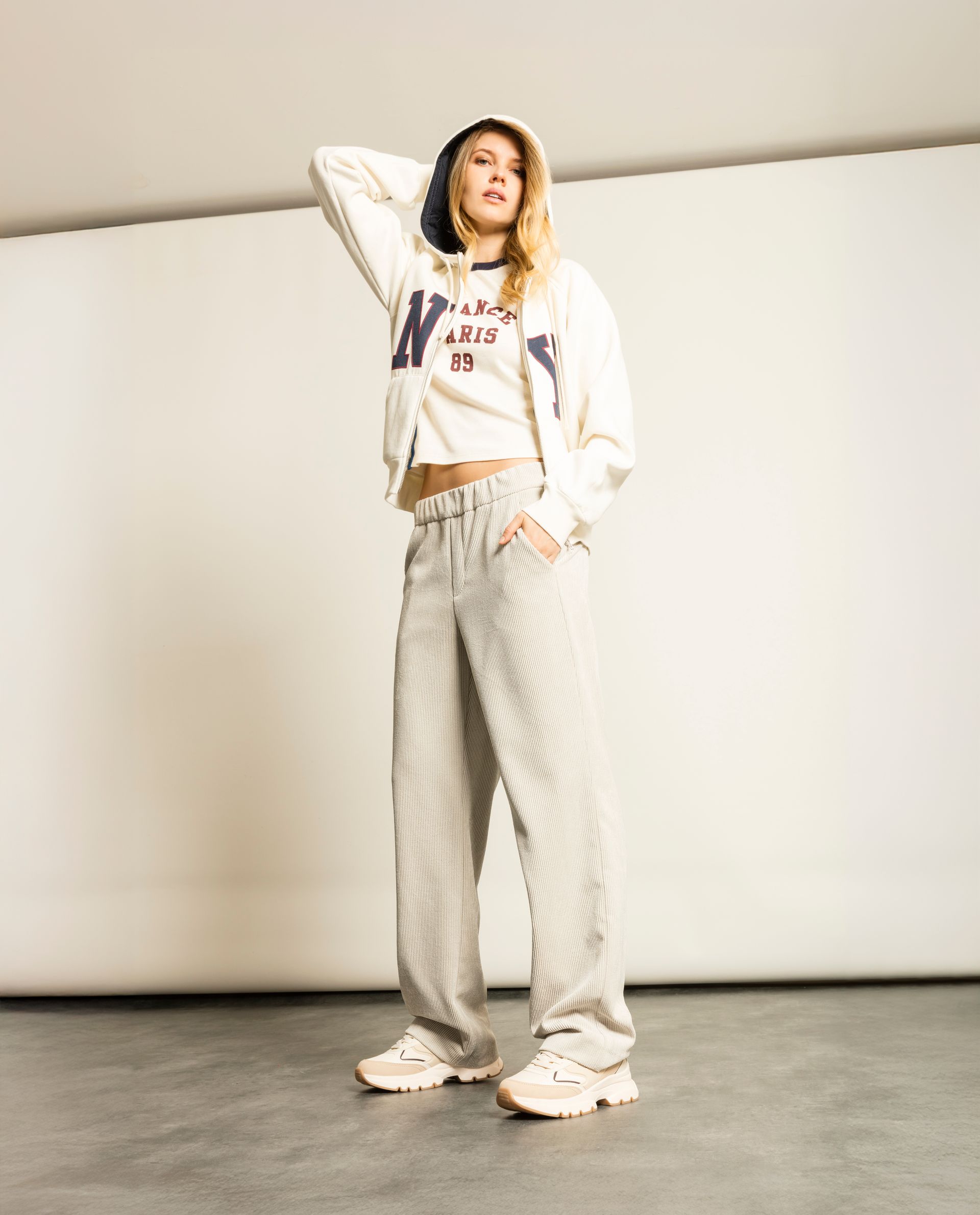 Frau in weißem Kapuzenpulli und gestreifter Hose, Hand am Kopf, posiert vor weißem Hintergrund.