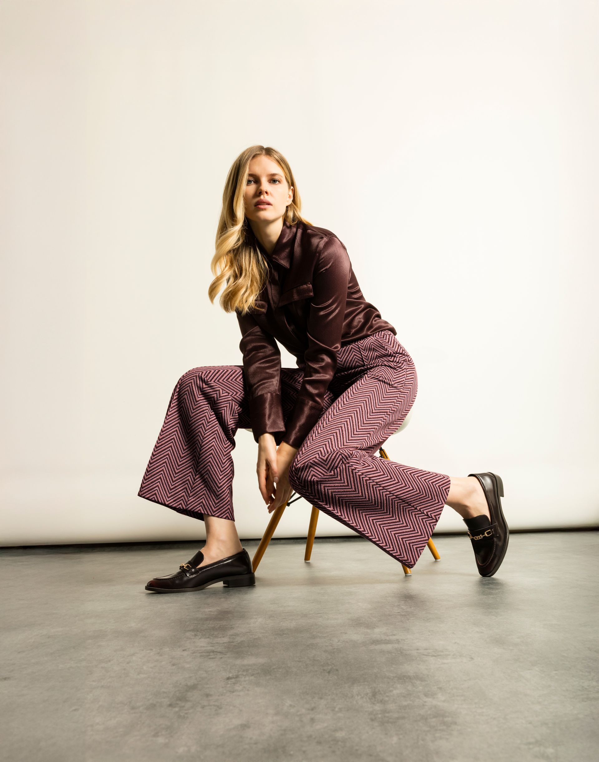 Eine Frau in bordeauxroter Kleidung kniet auf einem Hocker. Sie trägt ein Hemd mit Knöpfen, eine gemusterte Hose und Slipper.