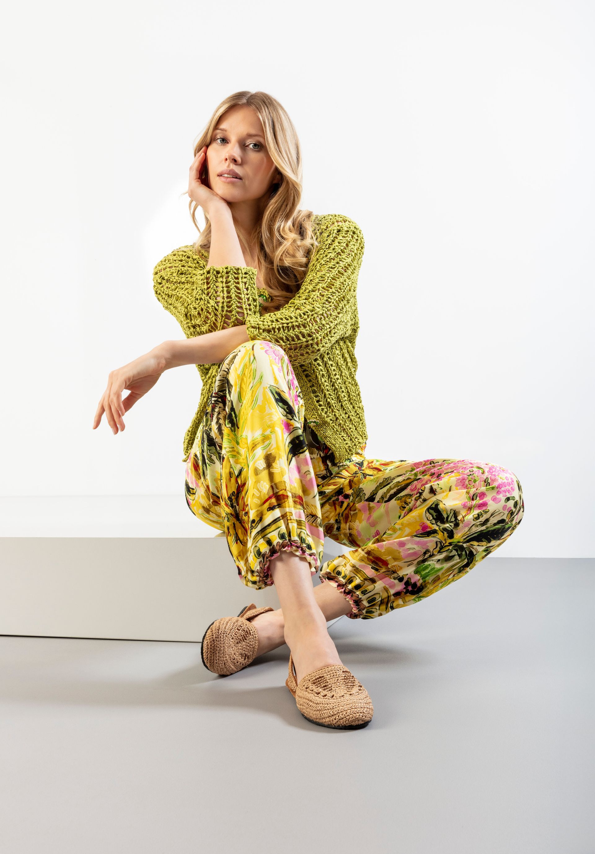 Frau in gelb gemusterter Kleidung, hockt auf einem weißen Block und schaut in die Kamera.
