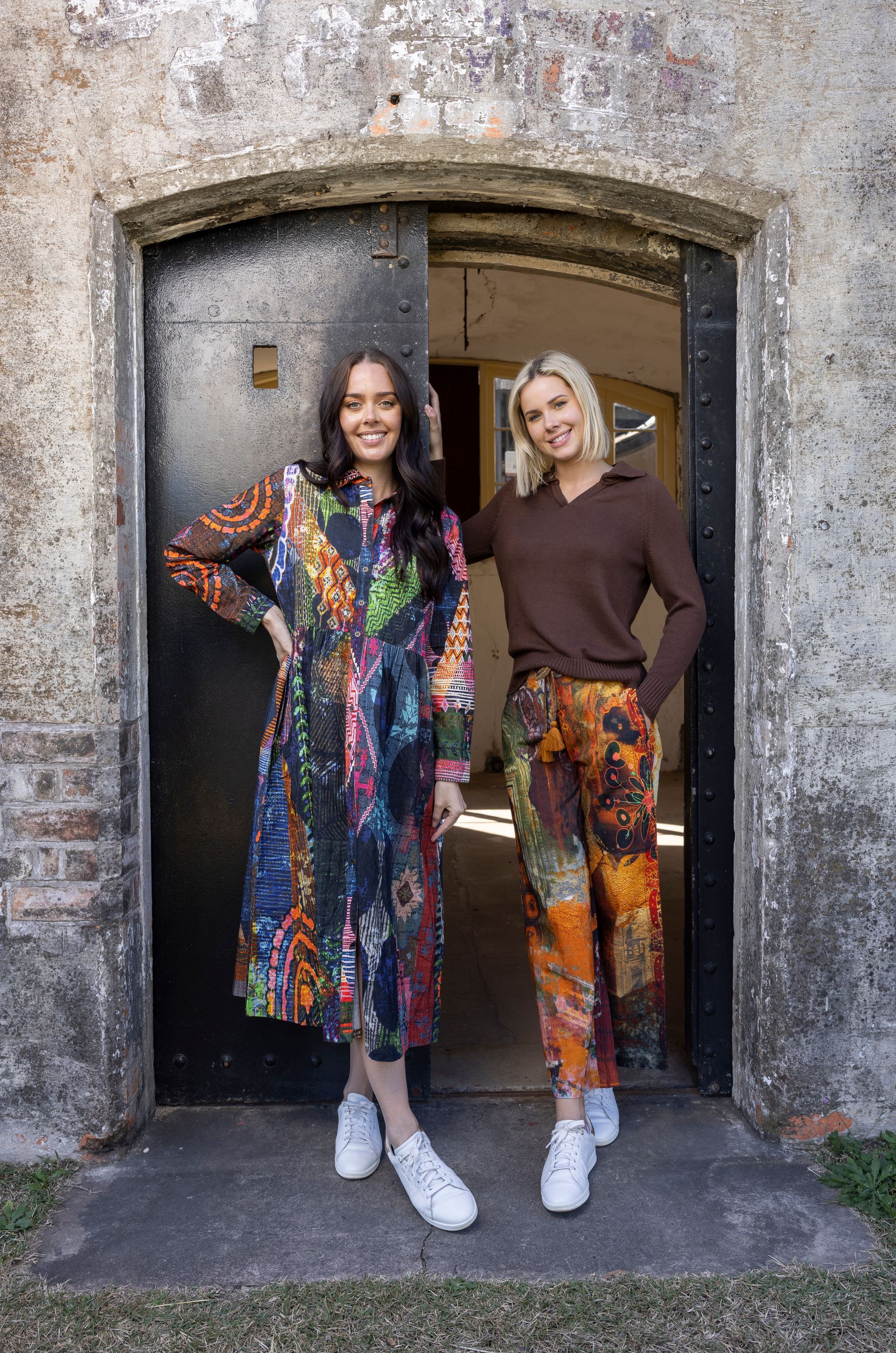 Zwei Frauen posieren in einem Türrahmen, die eine in einem farbenfrohen Kleid, die andere in einem braunen Pullover und einer gemusterten Hose.