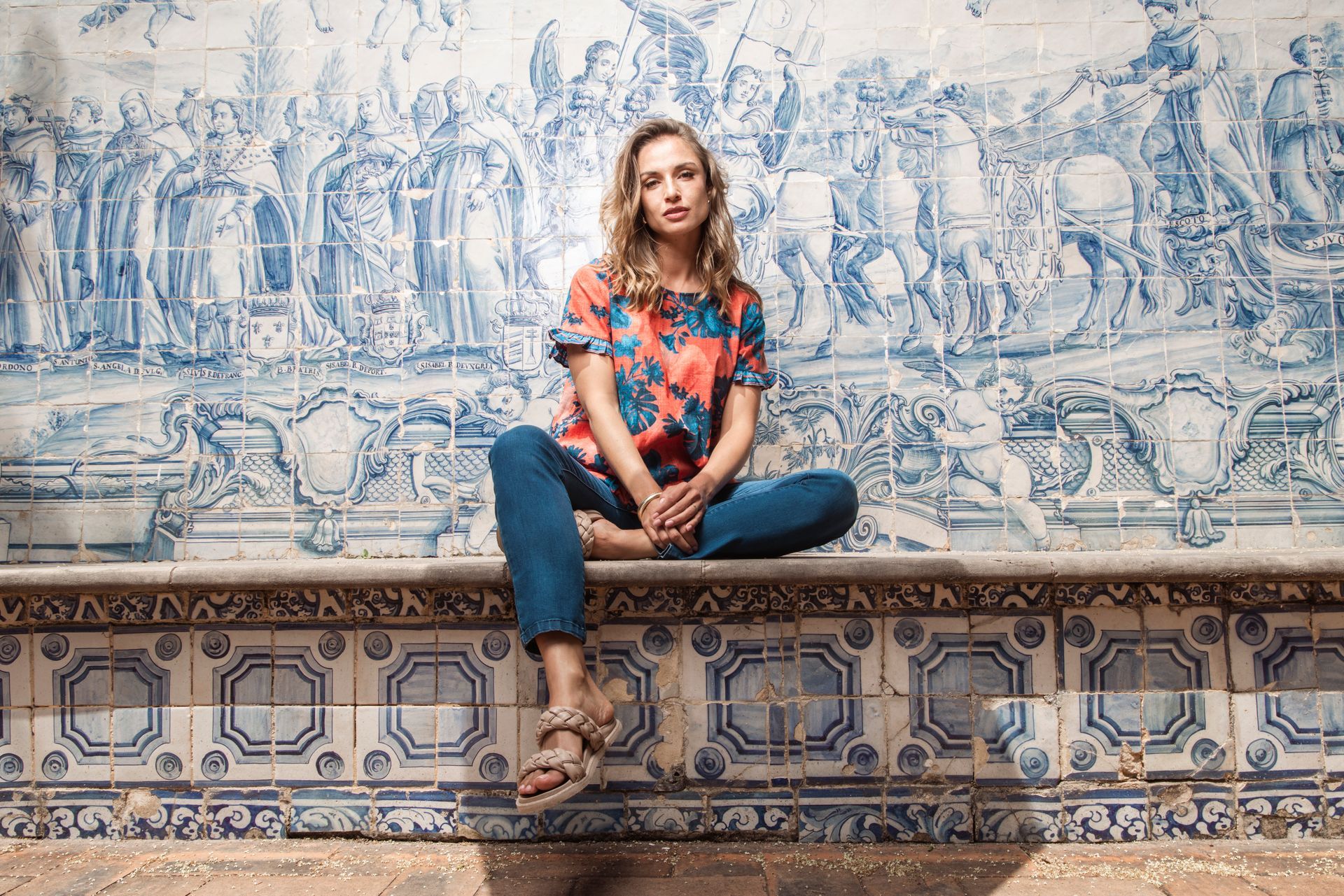 Eine Frau sitzt im Schneidersitz auf einer Bank vor einem blau-weißen Fliesenmosaik. Sie trägt ein geblümtes Oberteil und Jeans.