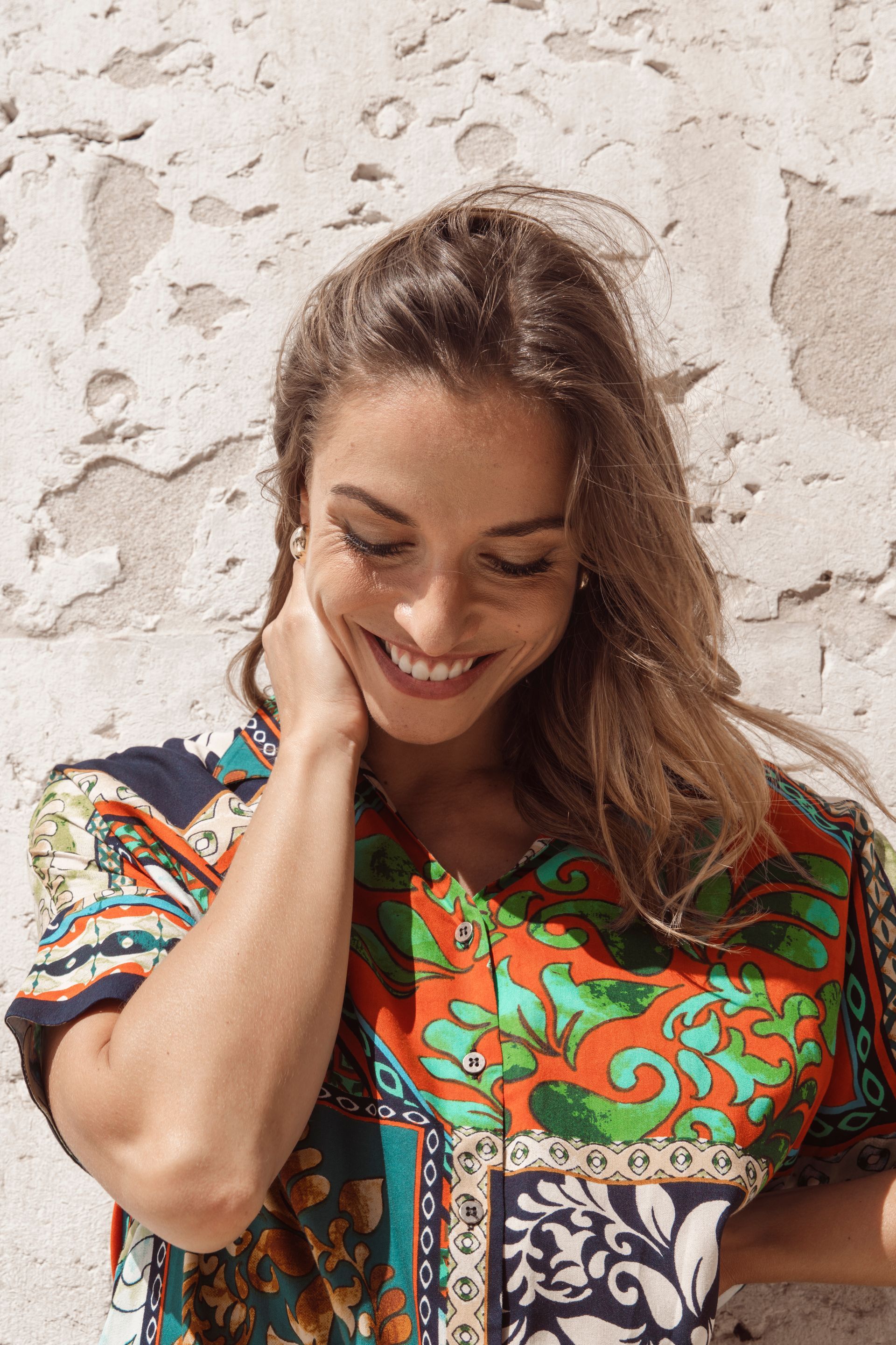 Frau mit langen braunen Haaren lächelt und trägt ein farbenfroh gemustertes Hemd vor einer weißen, strukturierten Wand.
