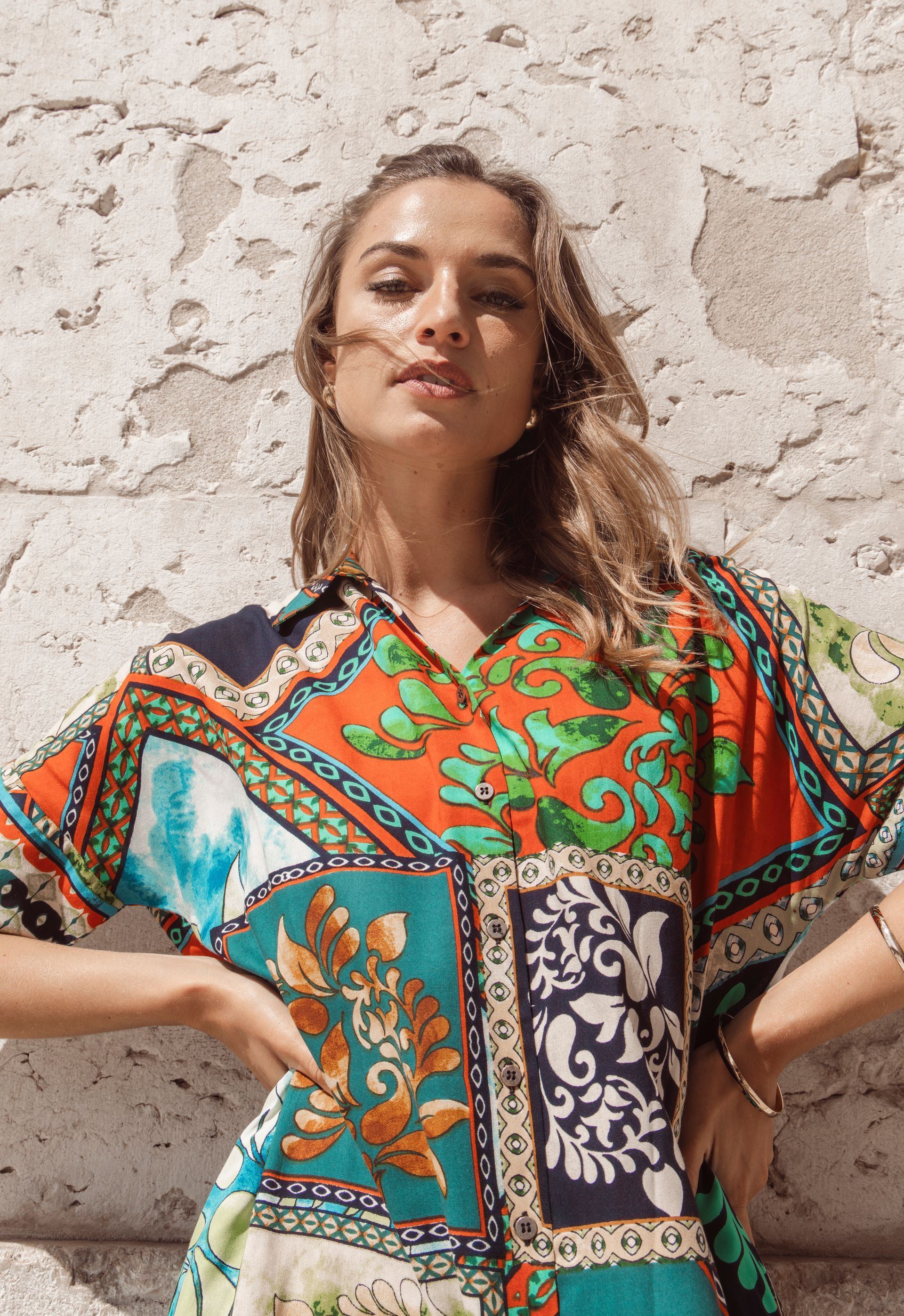 Frau in einem farbenfroh gemusterten Kleid, die Hände in die Hüften gestemmt, steht vor einer strukturierten Wand.