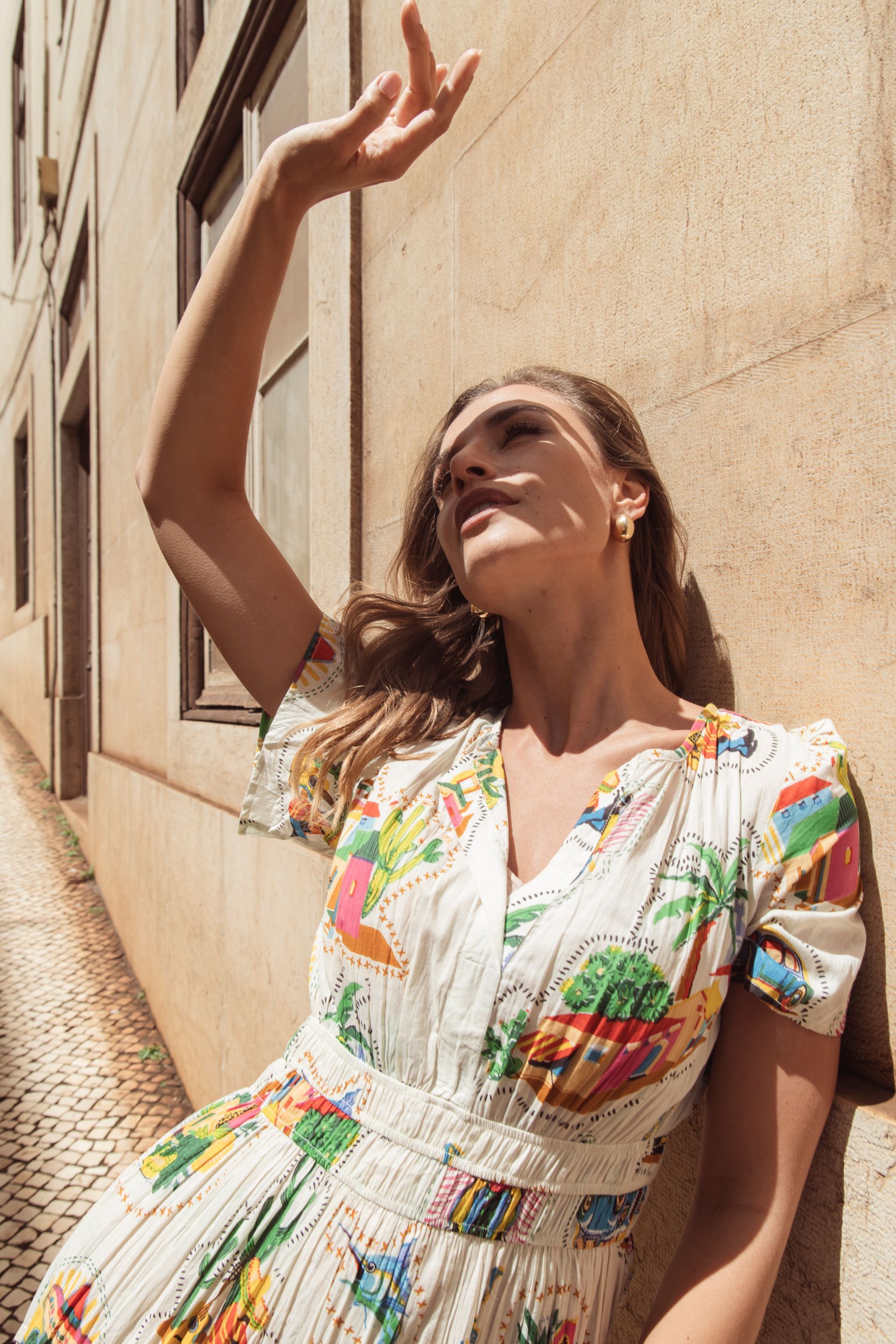 Frau in geblümtem Kleid, die ihre Augen vor der Sonne schützt, lehnt an einem sonnenbeschienenen Gebäude in einer Gasse.