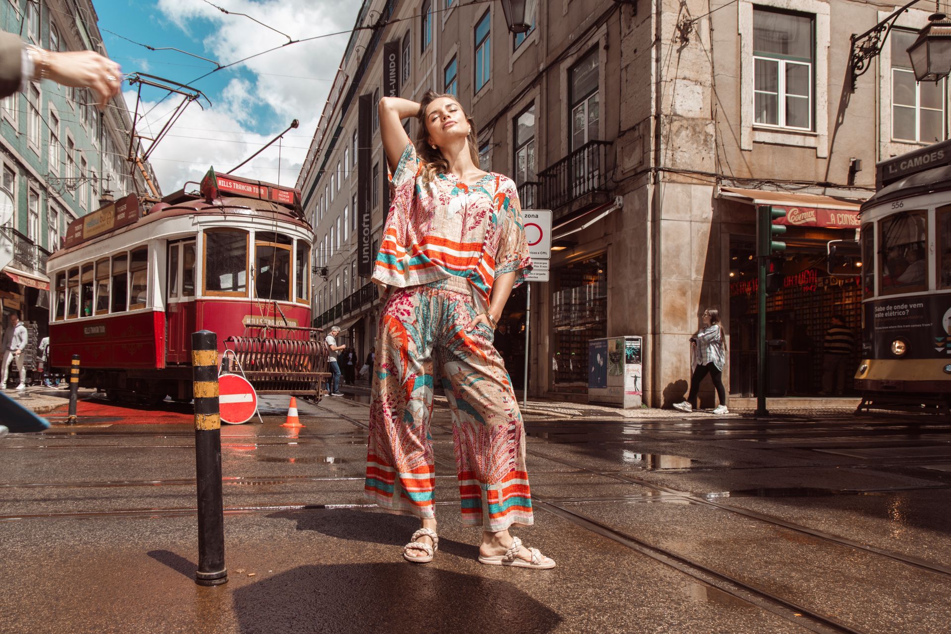 Frau in farbenfrohem Outfit posiert auf einer nassen Straße mit Straßenbahnen in Lissabon, Portugal.