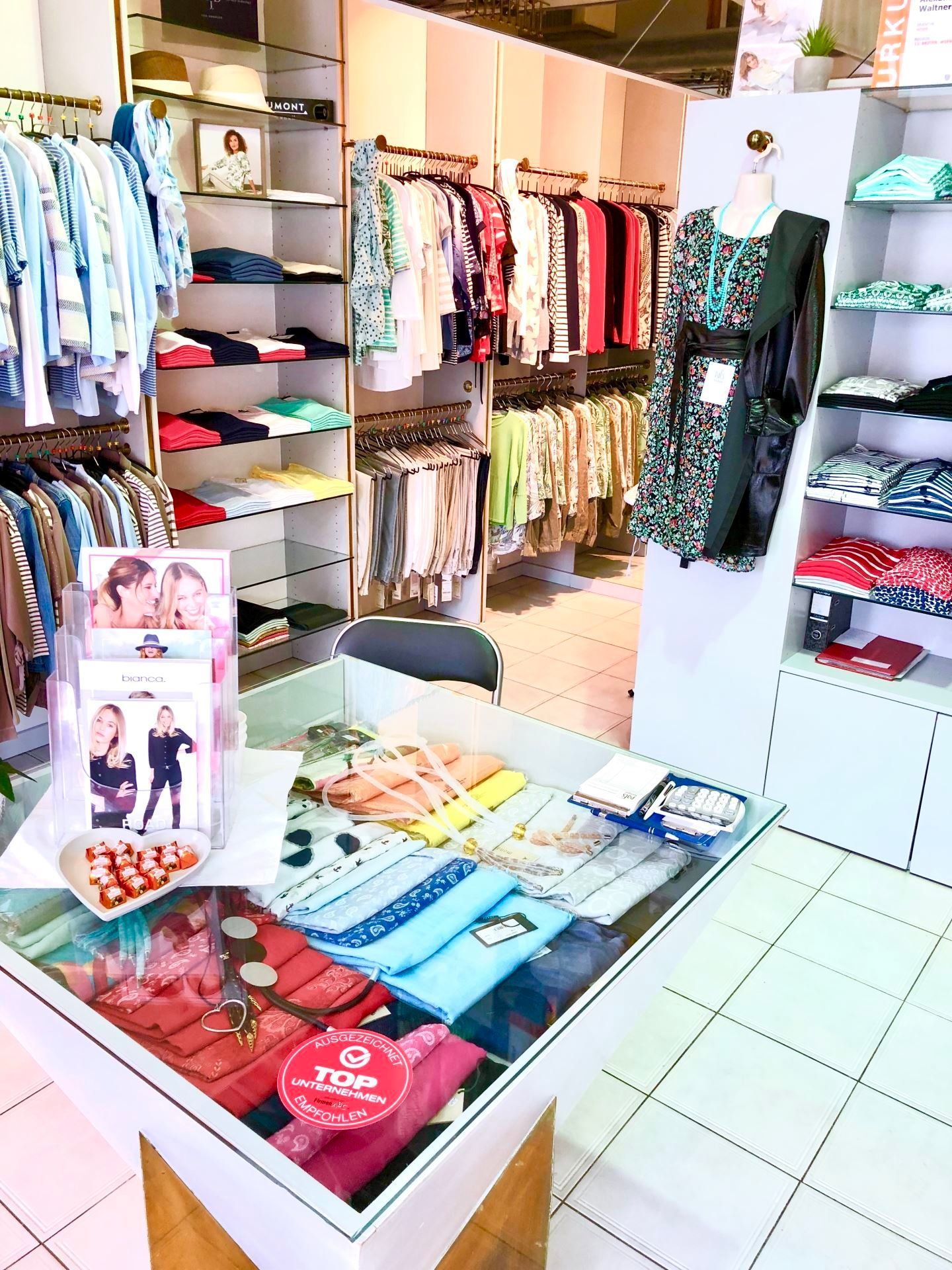 Innenansicht eines Bekleidungsgeschäfts mit Kleiderständern, einem Präsentationstisch mit Accessoires und einer Schaufensterpuppe.