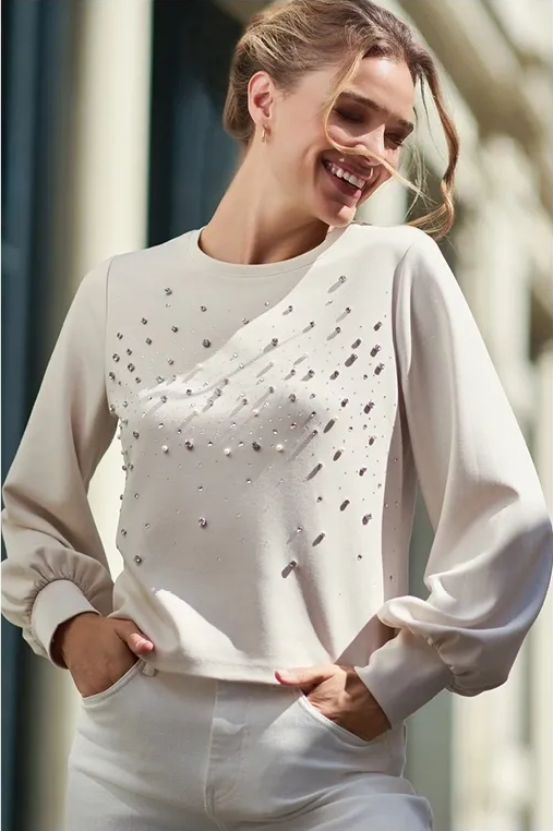 Frau in beigefarbener Bluse mit verzierter Vorderseite und weißer Hose lächelt im Freien.