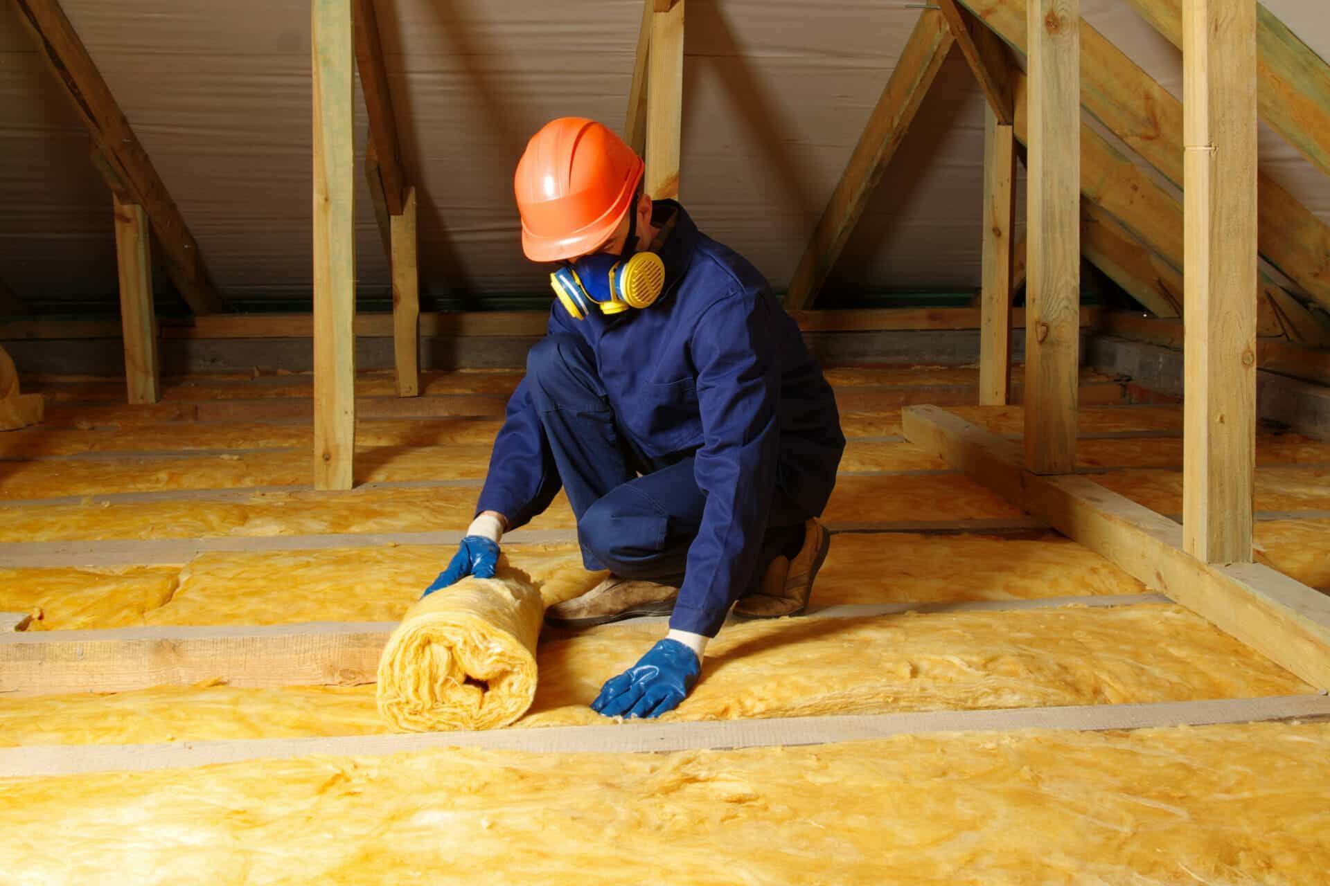 A man is kneeling down in an attic holding a roll of insulation.