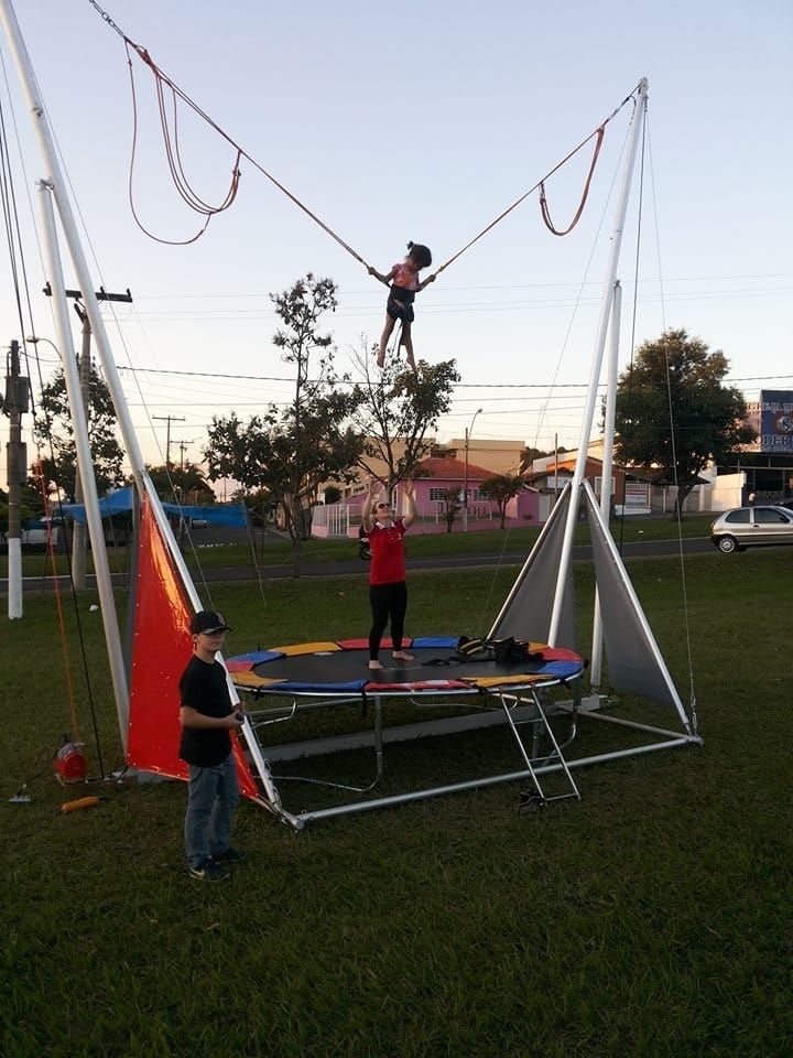 Pessoas em um trampolim de bungee jumping. Uma pessoa pulando, outra no trampolim, operador próximo. Campo de grama ao ar livre.