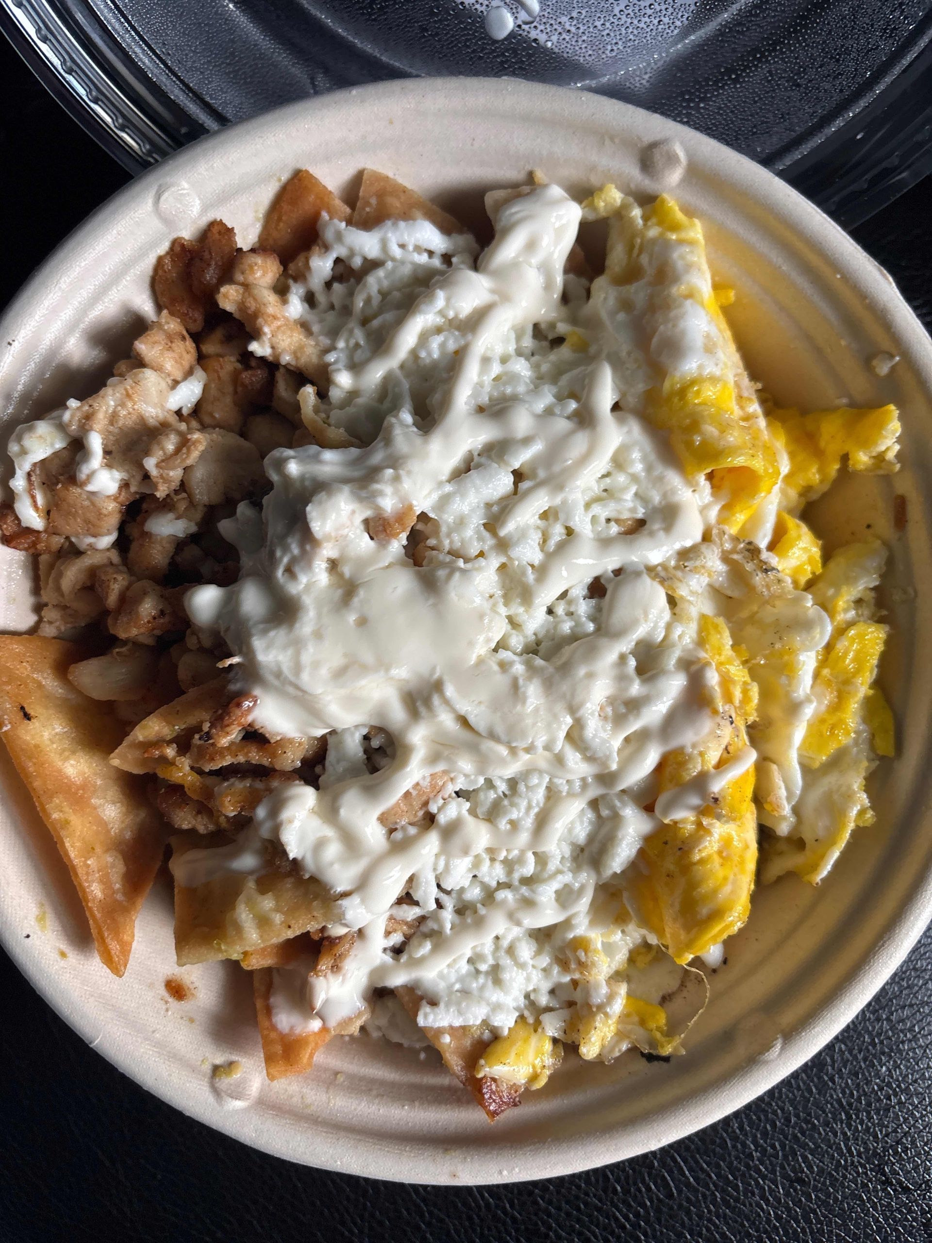 A bowl of chilaquiles featuring fried tortilla chips, scrambled eggs, and chicken topped with a white creamy sauce.