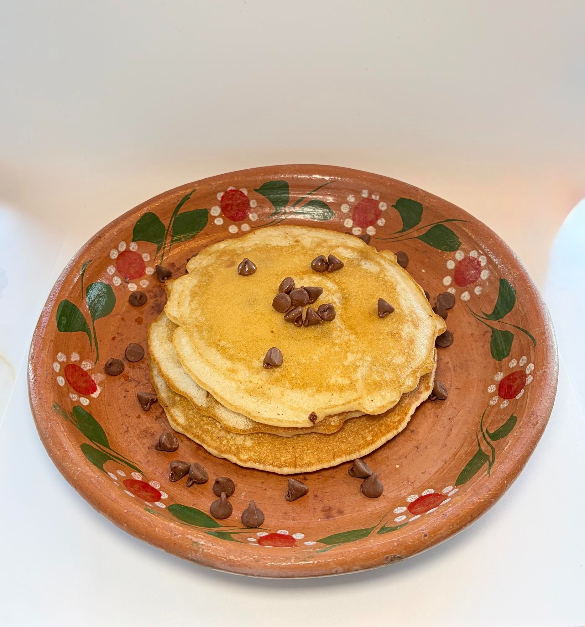 A stack of pancakes topped with chocolate chips sits on a decorated terracotta plate against a white background.