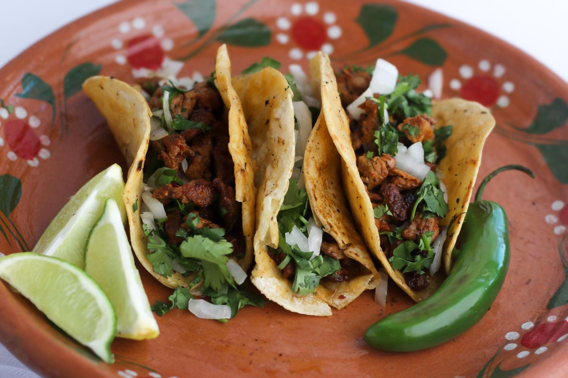 Three corn tortilla tacos topped with meat, onion, and cilantro on a floral clay plate with lime wedges and a chili.