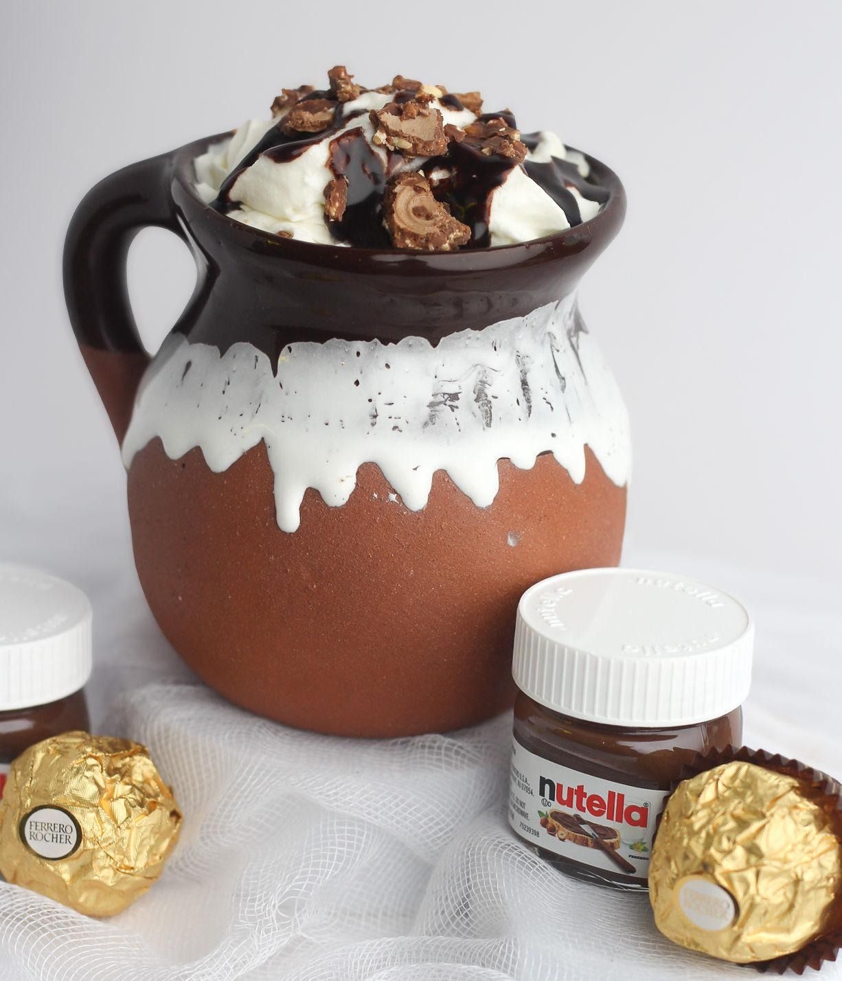 A clay mug filled with whipped cream, chocolate sauce, and candies, next to small Nutella jars and Ferrero Rocher candies.