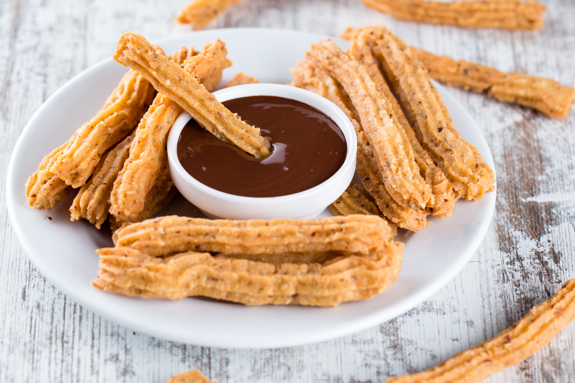 A plate of golden-brown churros surrounding a small bowl of dark chocolate dipping sauce on a white wooden surface.