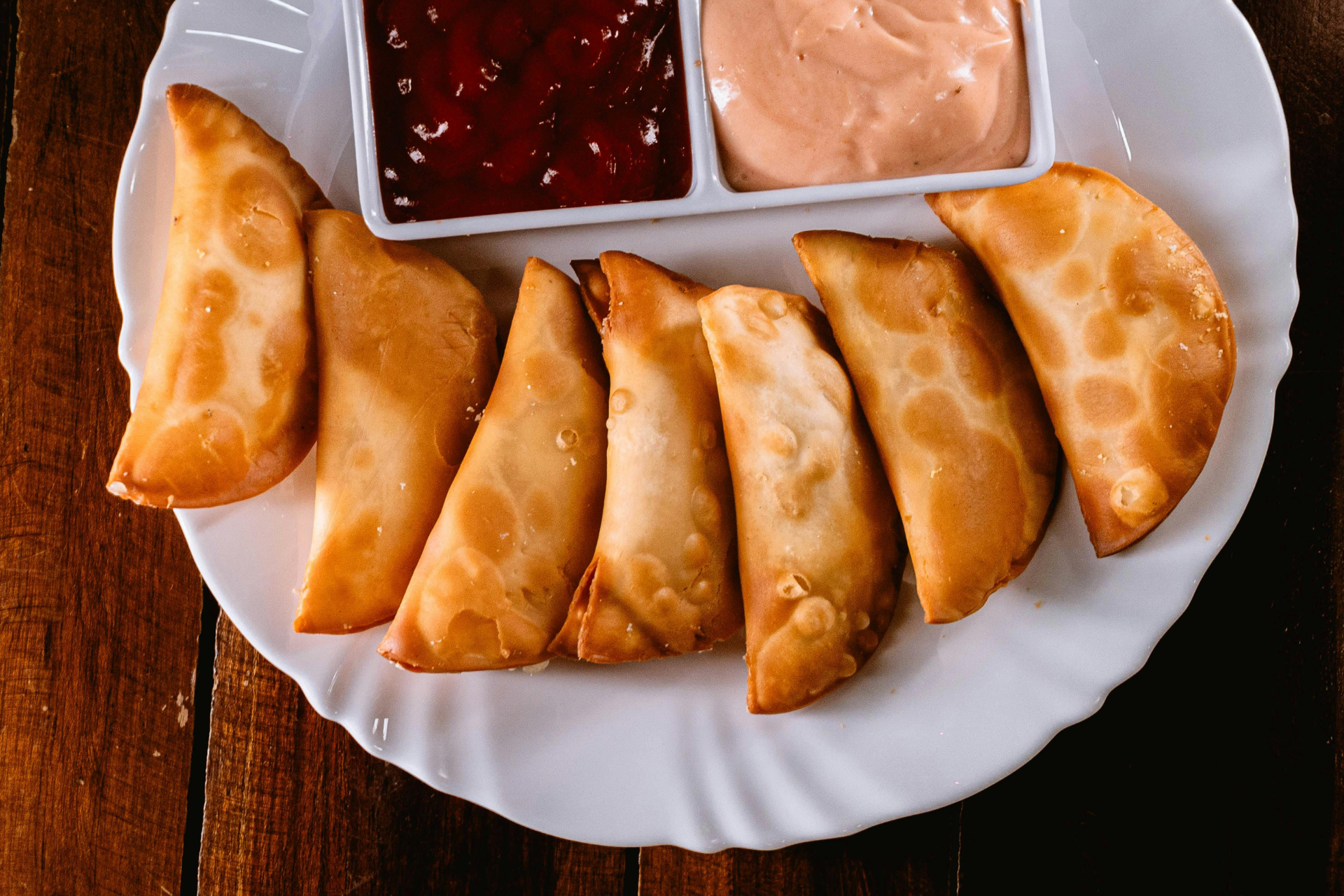 A white plate holds several golden-brown fried empanadas and two small dishes of red and creamy dipping sauces.