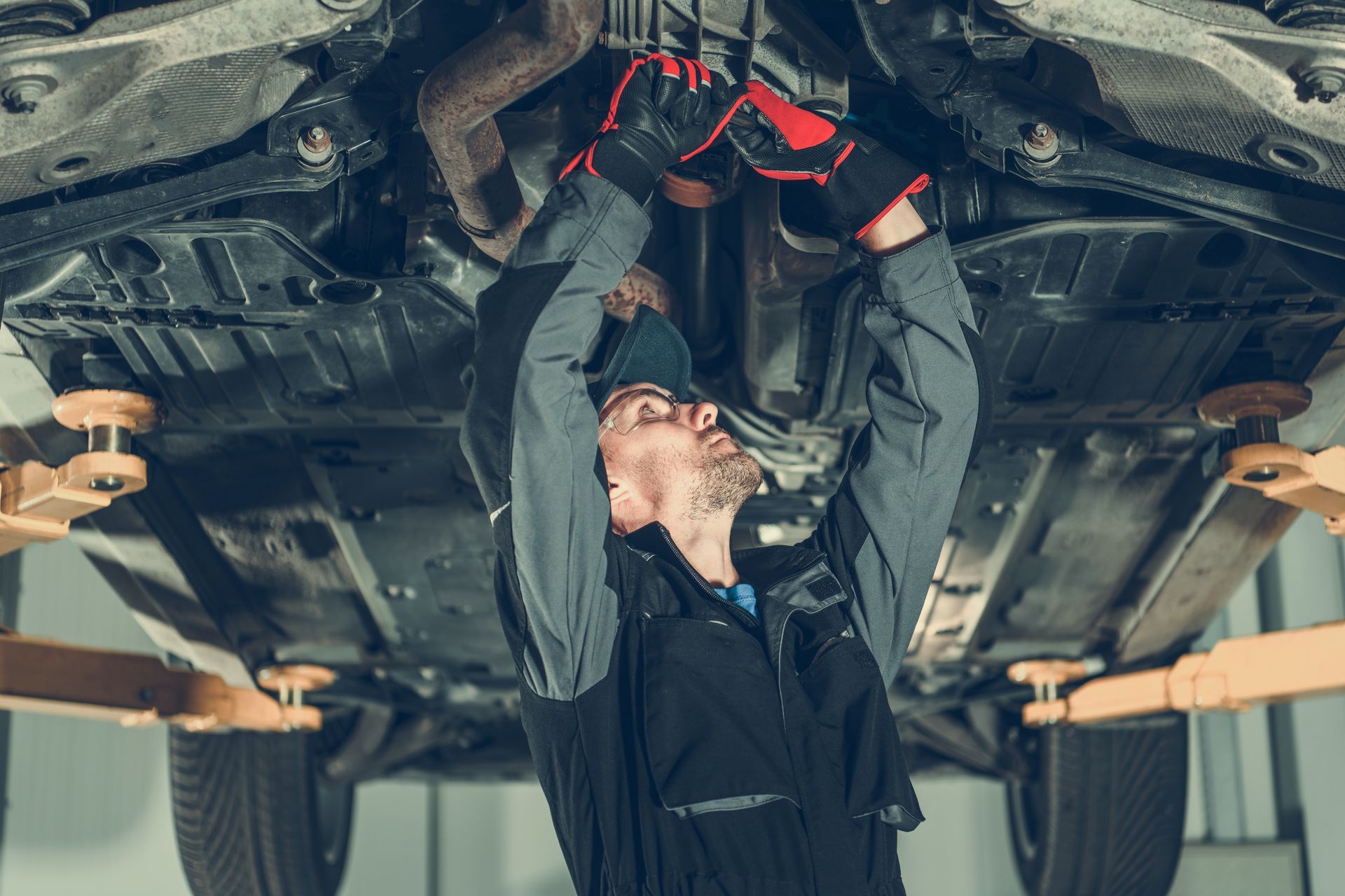 Mechanic works under a car lifted on a hoist. He wears gloves and a cap. Workshop setting.