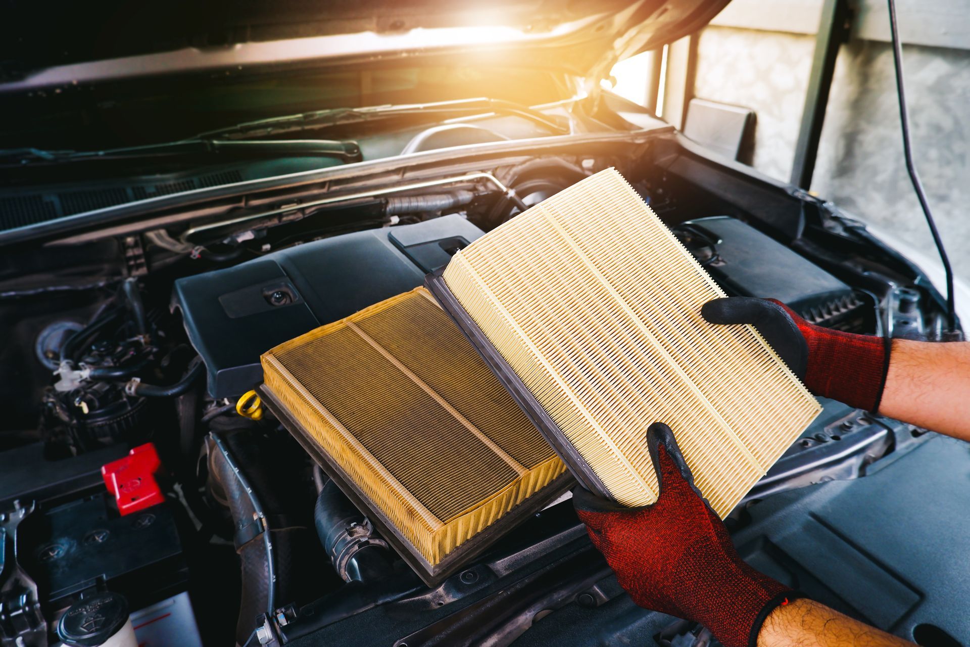 Hands in gloves holding a new and old air filter next to a car engine.