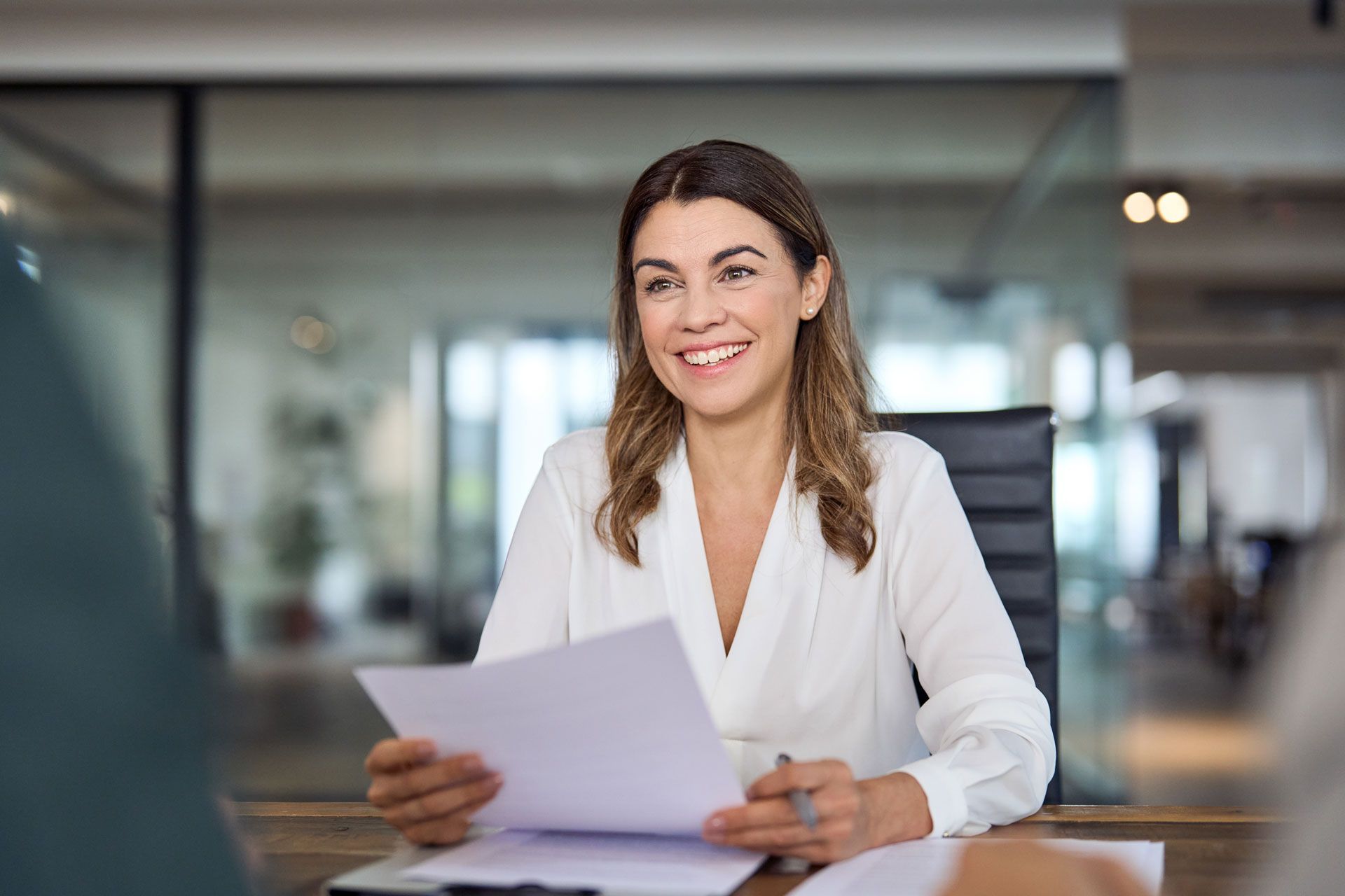 Woman in white shirt smiles, holding papers, at a desk in an office.