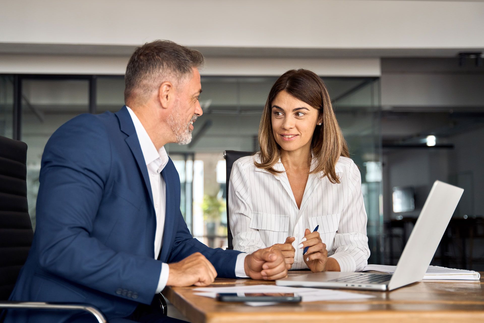 Man and woman in an office, discussing work at a desk with a laptop and documents.