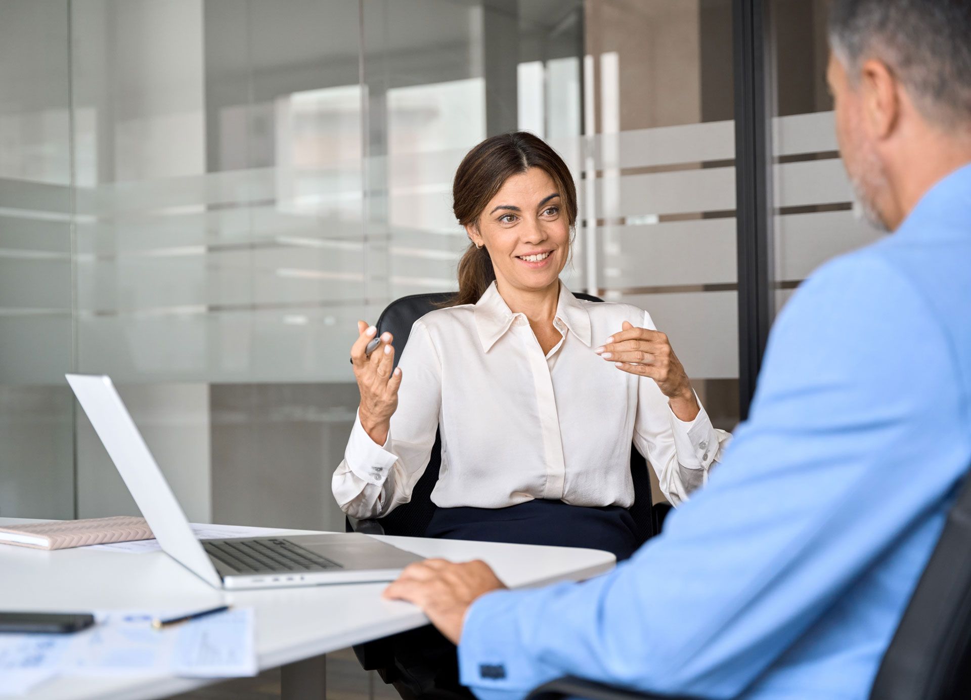 Woman in white shirt gestures, talking to person at a desk in a modern office.