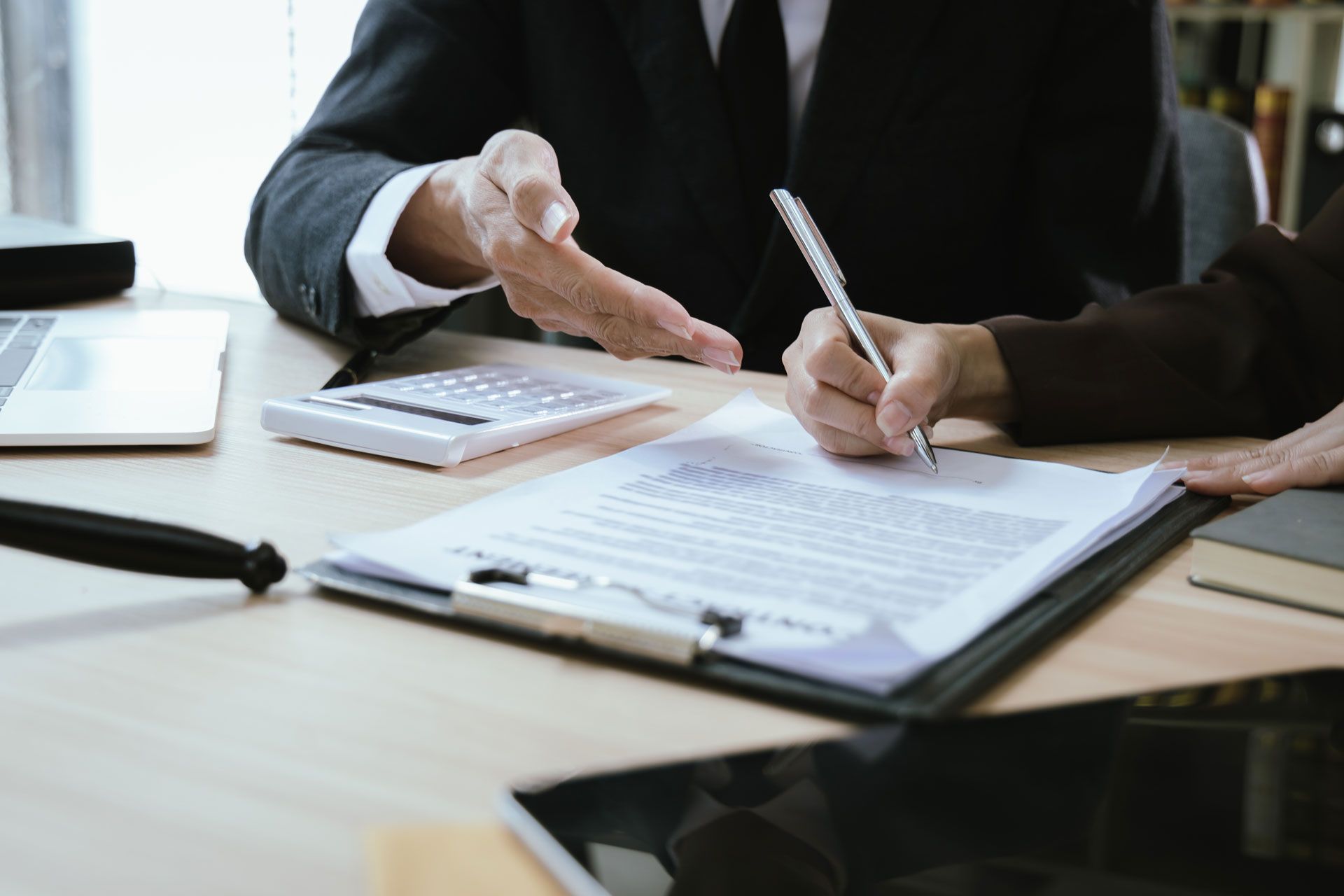 Person in suit points to document while another signs at a desk with calculator and pen.