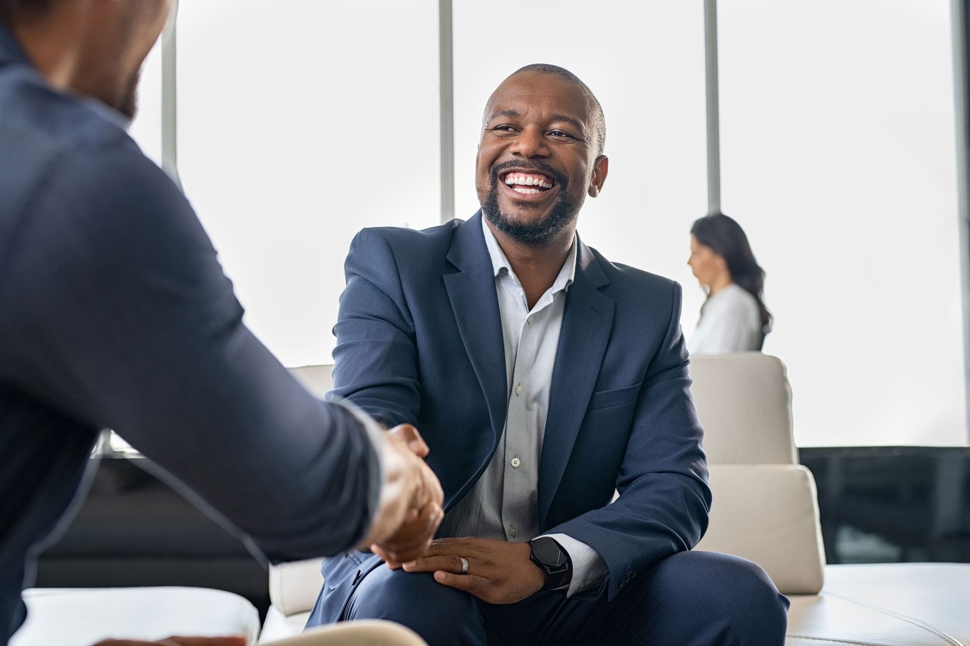 Man in suit smiles shaking hands with another person; office setting.
