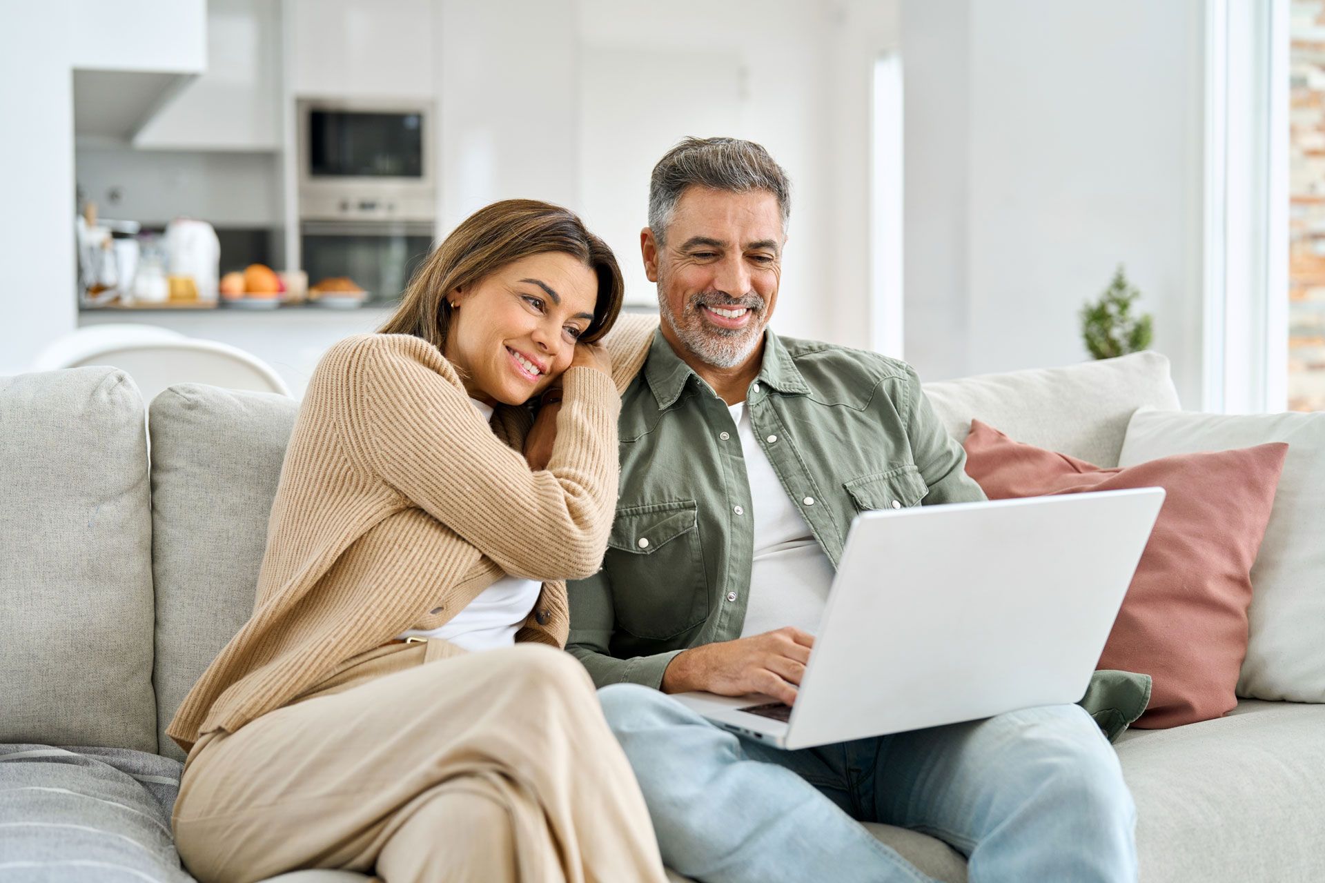 Couple on sofa looking at laptop, smiling. Neutral tones, indoor setting.