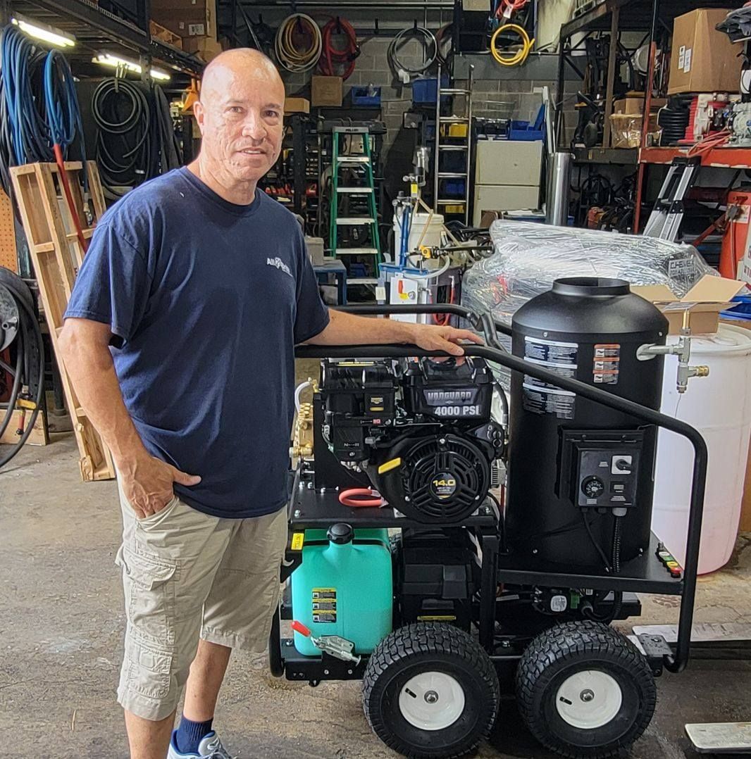 Man stands next to a black and silver pressure washer with a large tank and wheels. Inside a workshop.