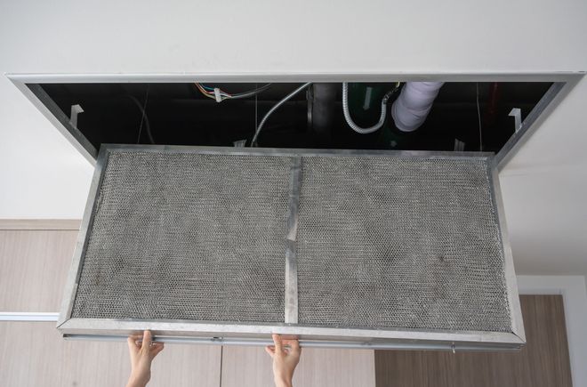 Hands holding a large air filter panel removed from a ceiling vent.