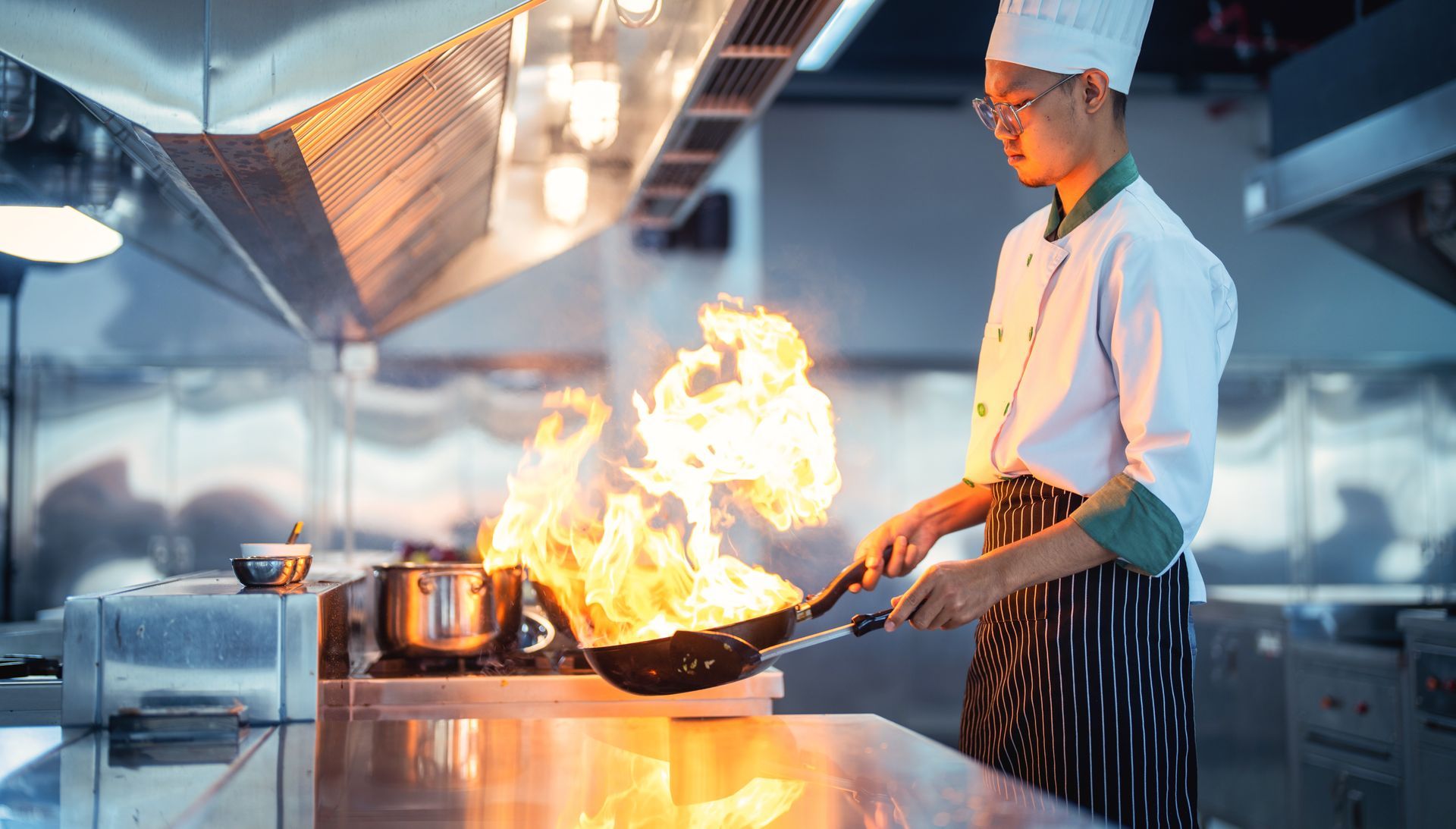 Chef in a kitchen, holding a pan with flames rising, cooking on a stovetop.