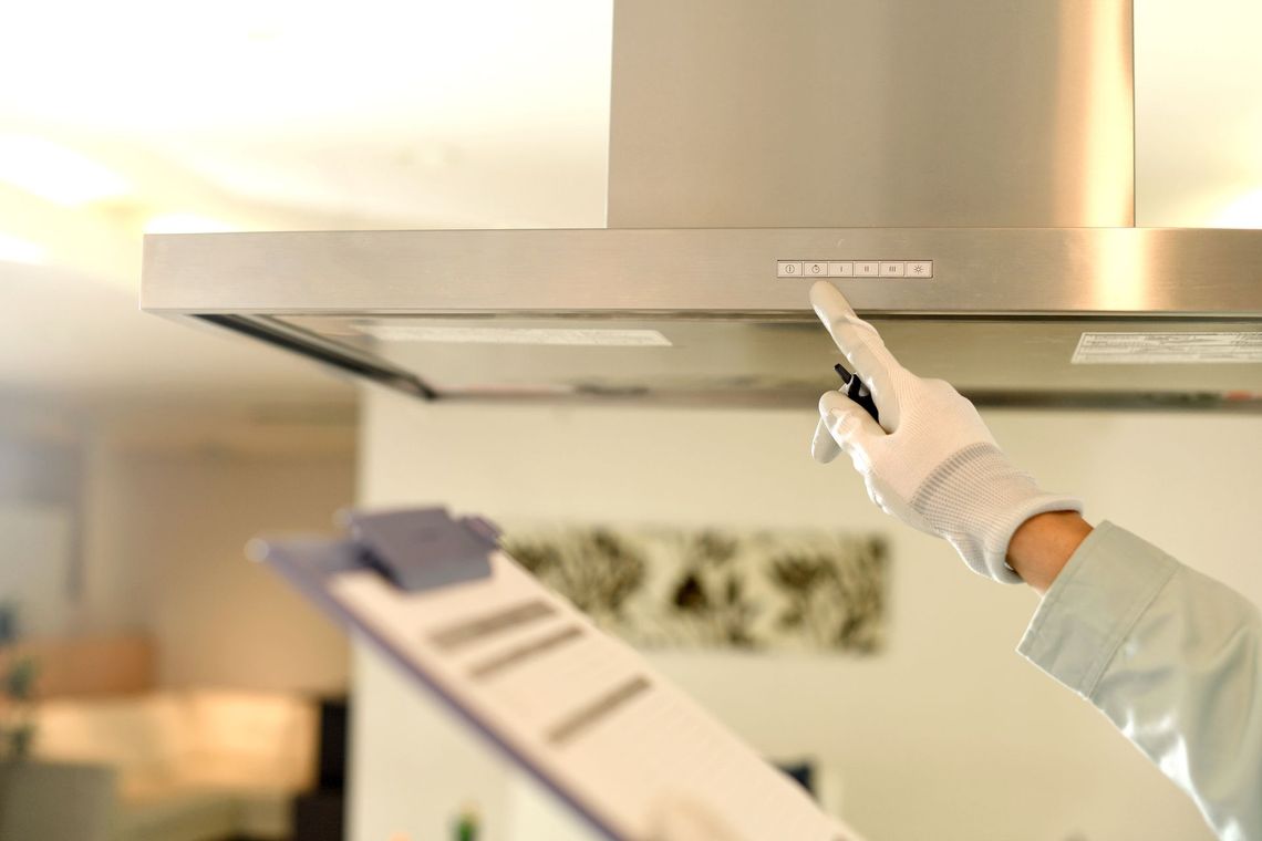 Person in white gloves pointing at a range hood control panel, holding clipboard in a kitchen.