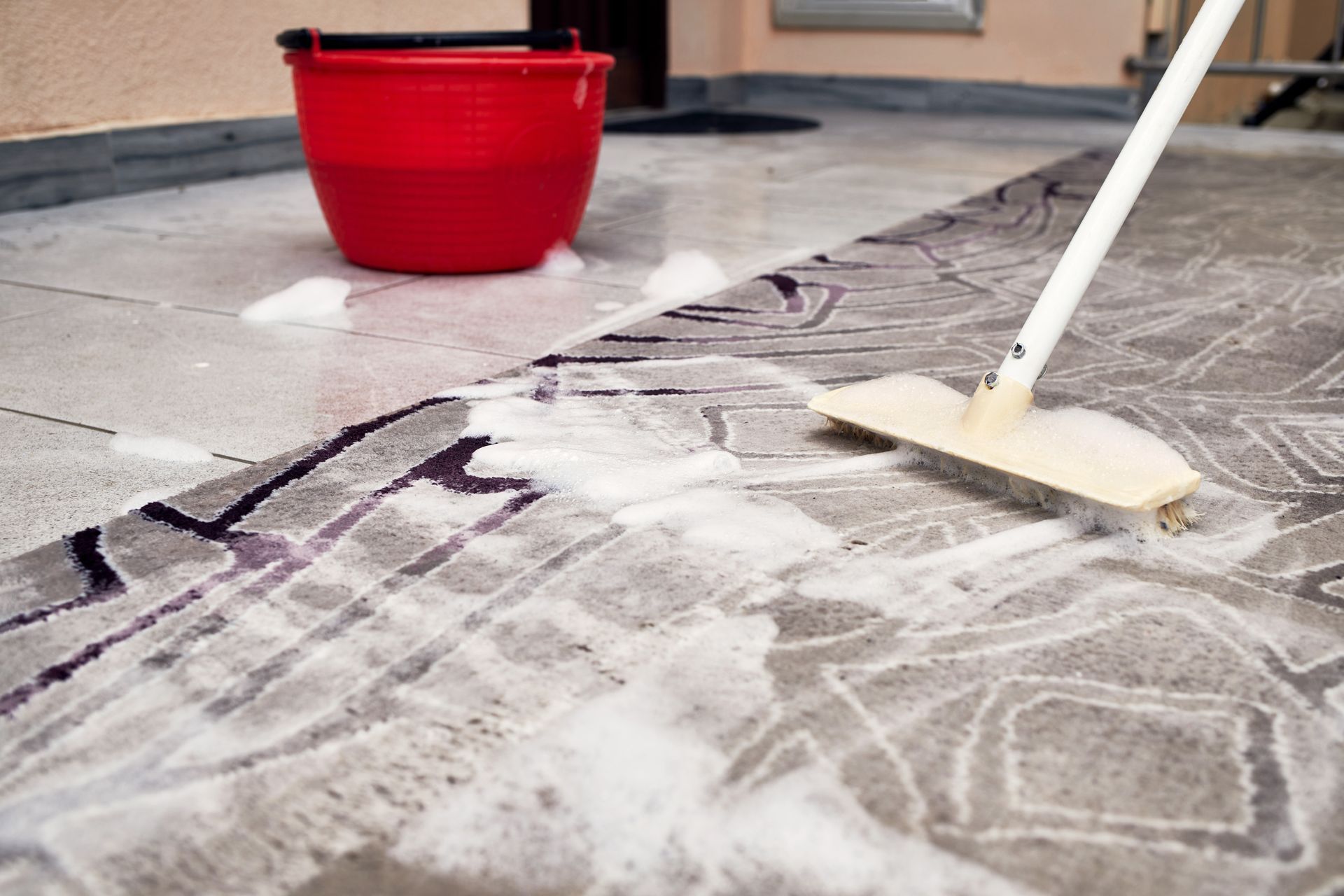 Red bucket of soapy water next to a rug being cleaned with a mop on a tiled floor.