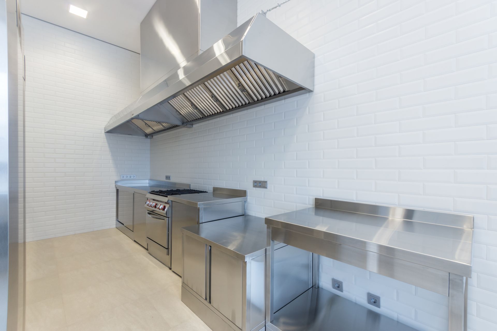 Stainless steel commercial kitchen with vent hood, stove, and prep tables. White tiled walls.