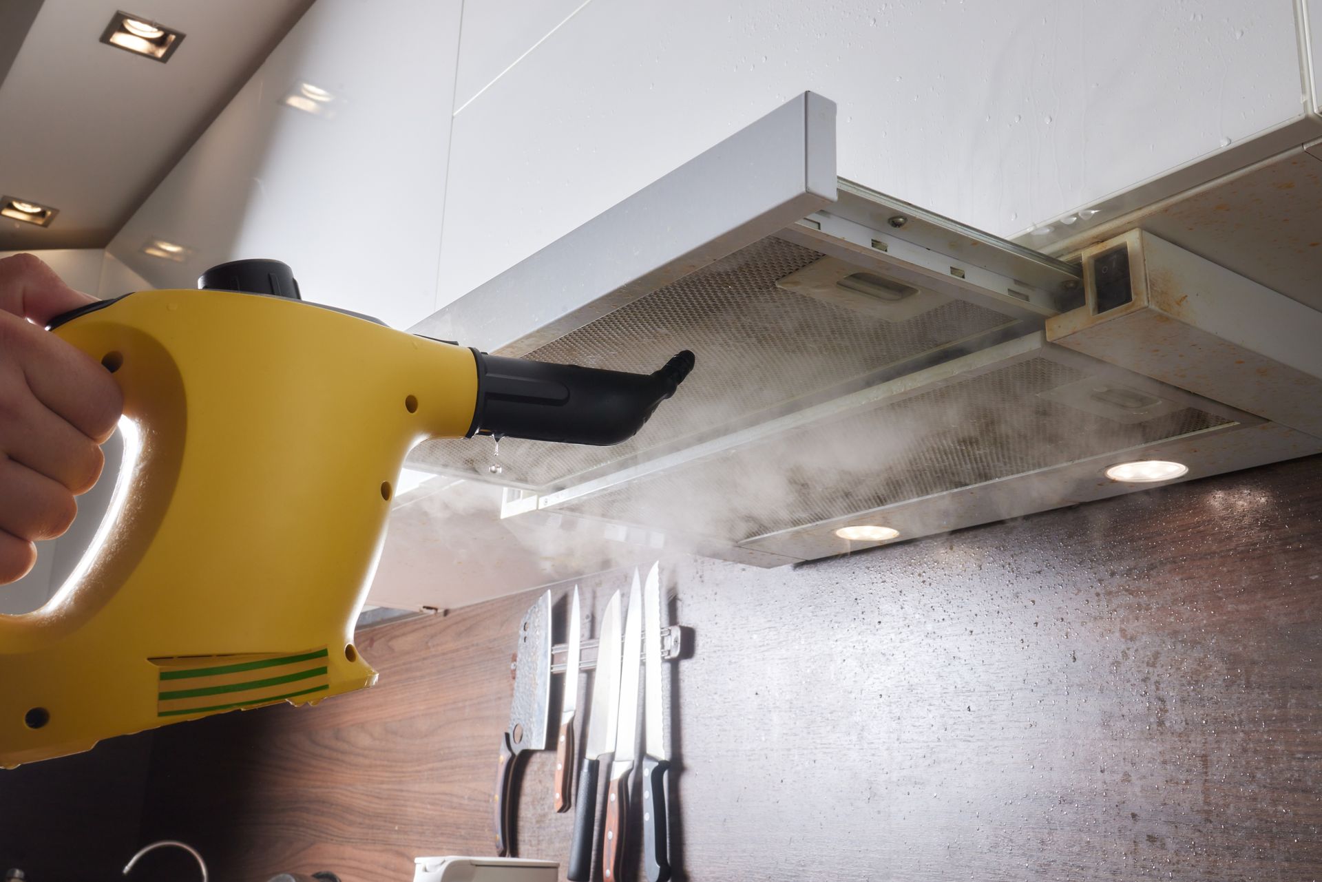A person uses a yellow steam cleaner to clean a kitchen range hood.
