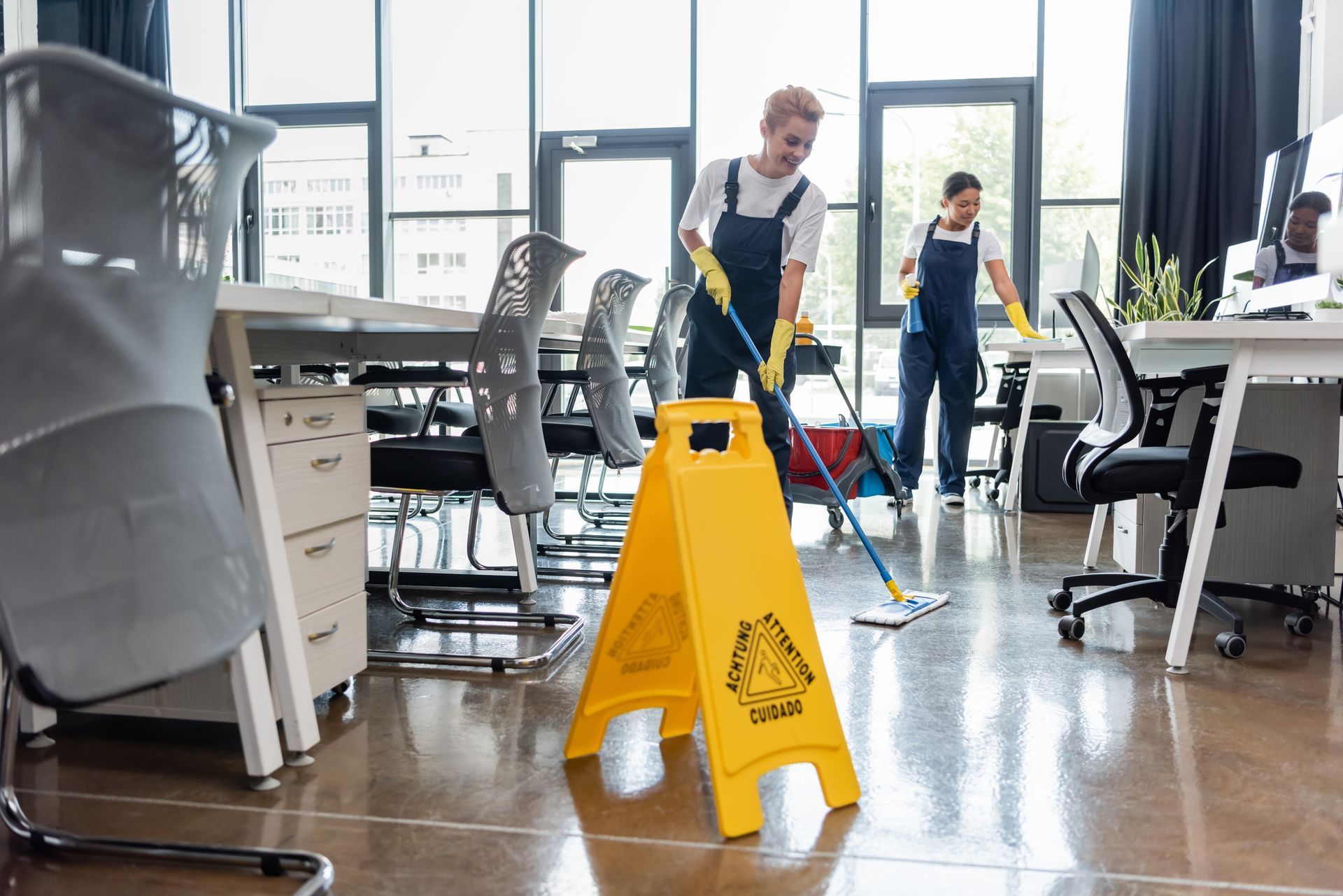 Two people cleaning an office floor with mops, a yellow wet floor sign is in the foreground.