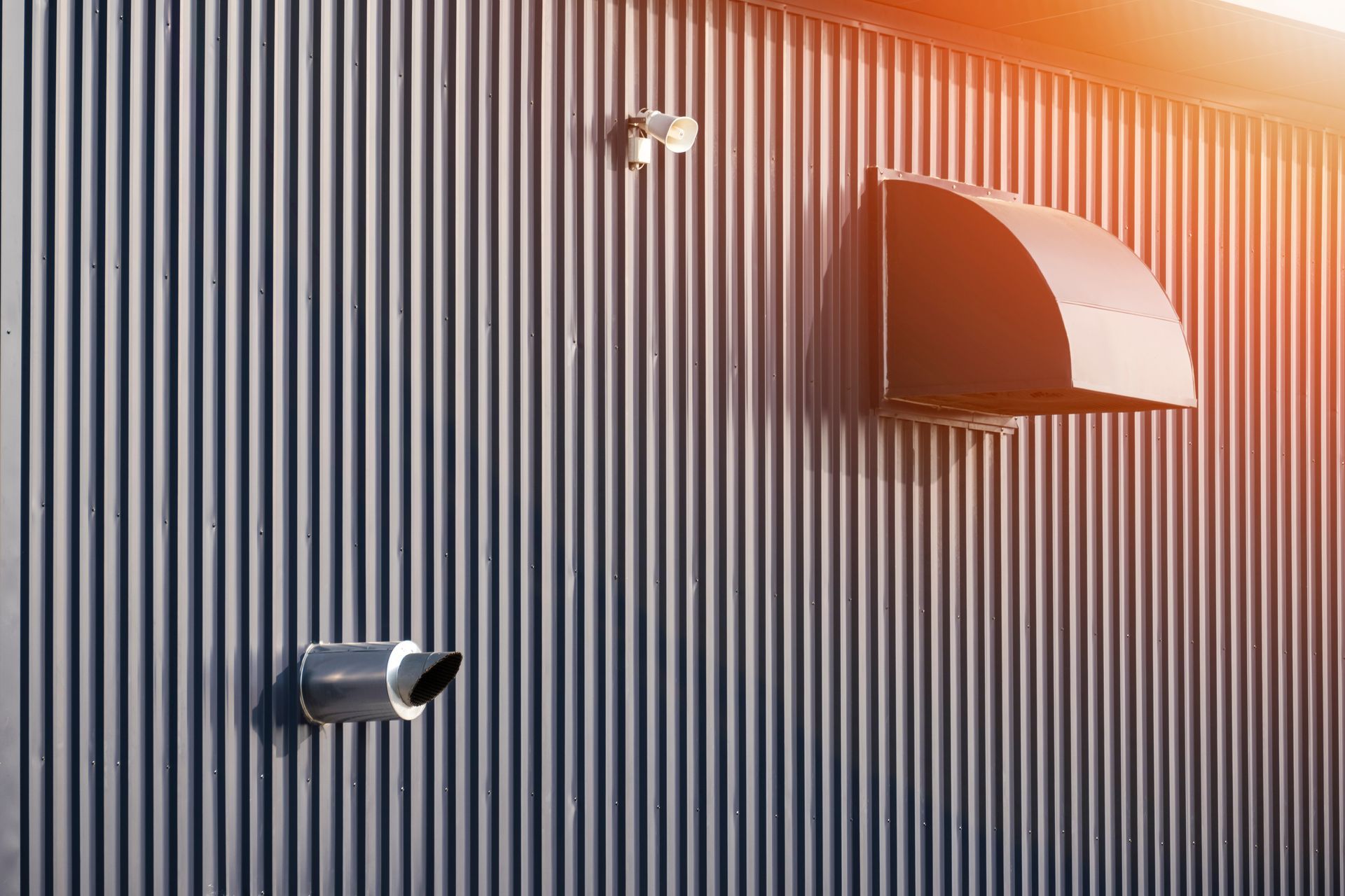 Gray corrugated metal wall with exhaust vent, pipe, and security cameras. Bright sunlight.