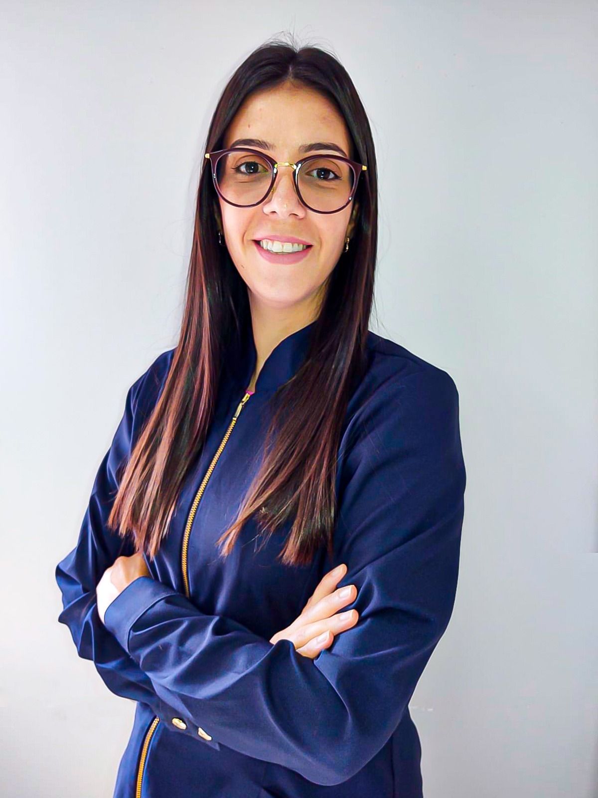 Mulher de uniforme azul, óculos, braços cruzados, sorrindo, dentro de casa.