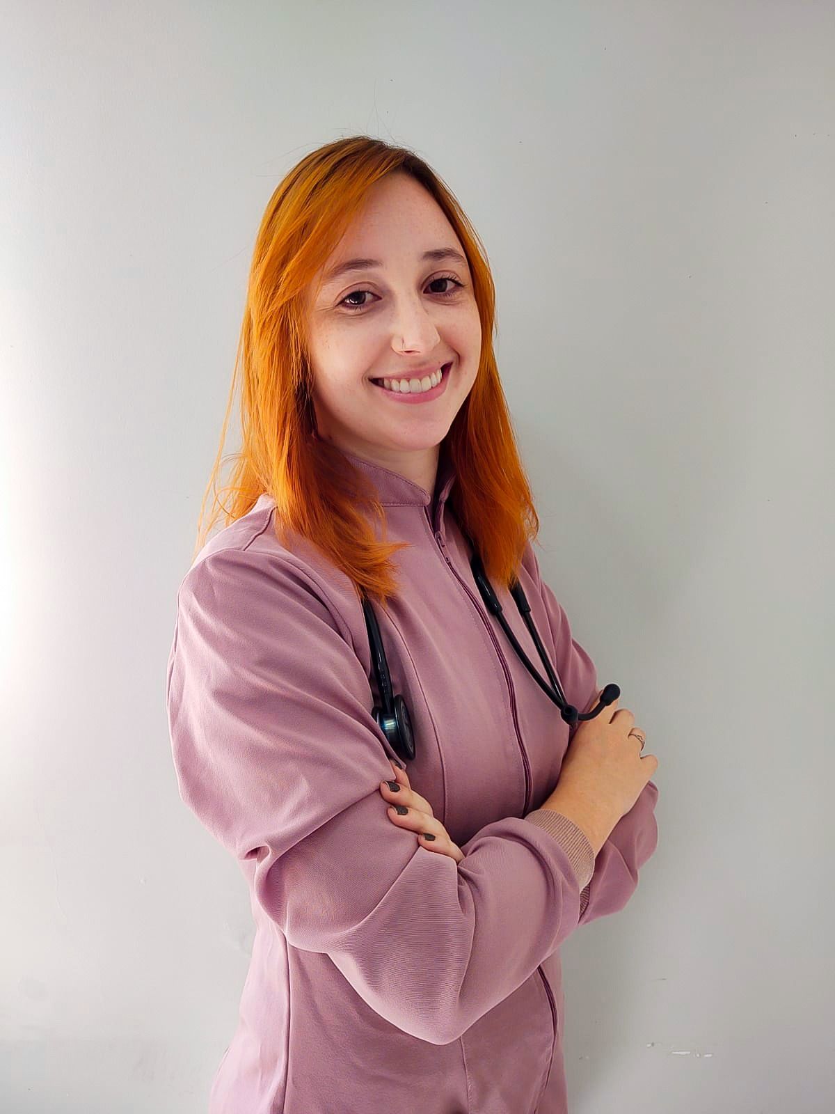Mulher com cabelos ruivos, vestindo um uniforme rosa, sorrindo, segurando um estetoscópio.