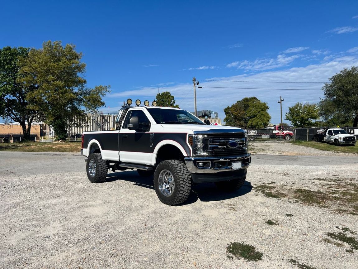 White and black lifted pickup truck parked on gravel under a blue sky. | Justin's Exhausted